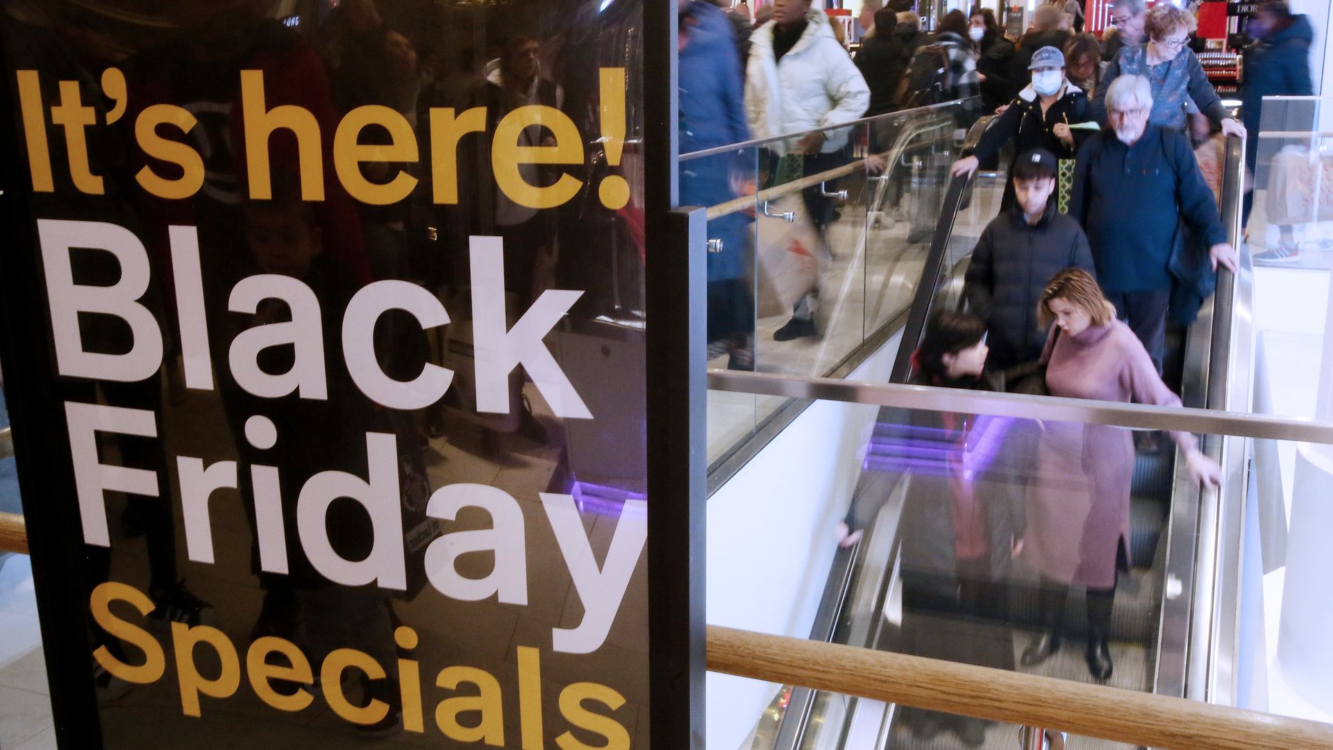 A sign that reads "It's here! Black Friday special" in front of shoppers on an escalator 
