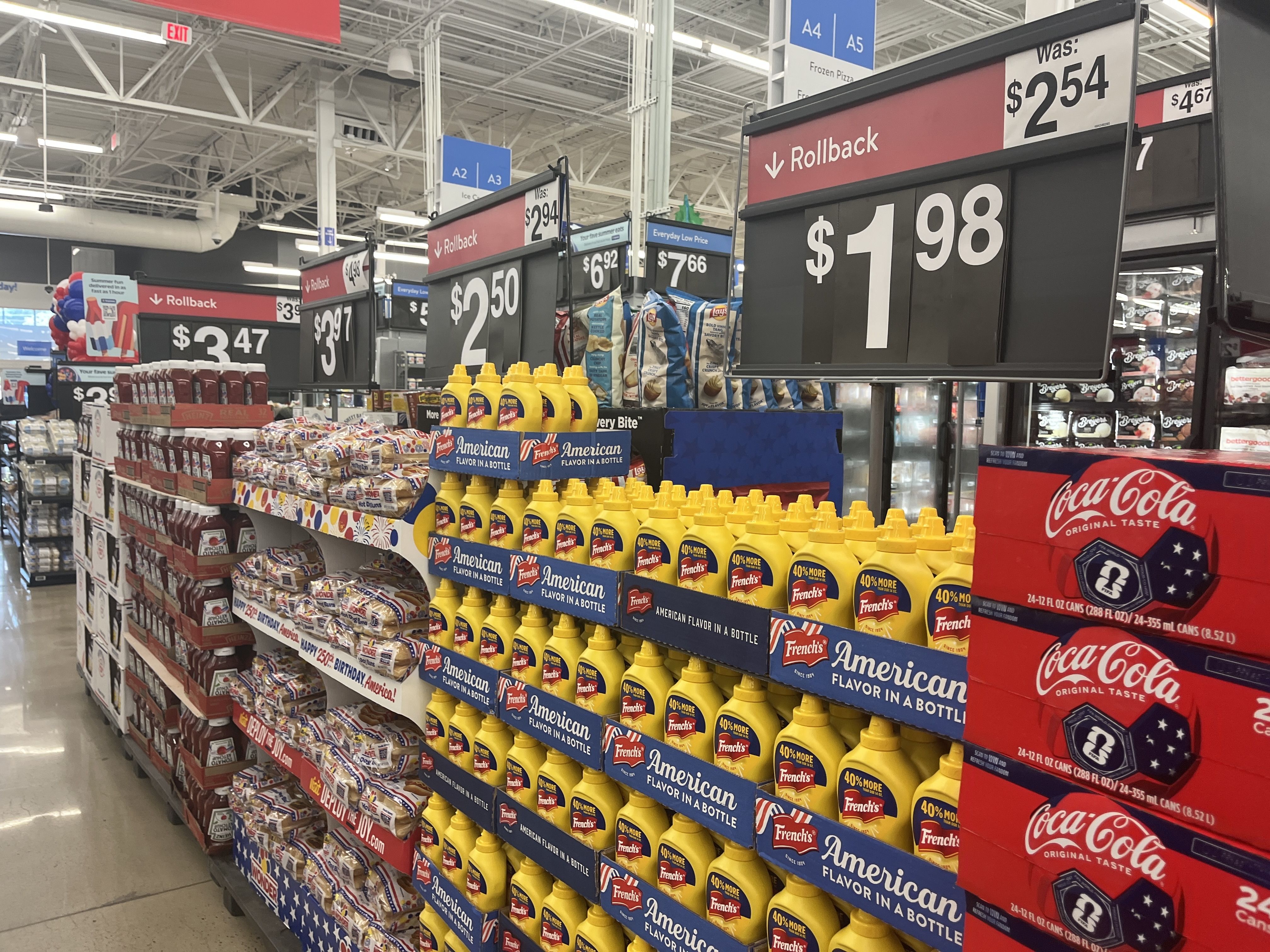 Inside a bright supermarket aisle with red Rollback signs and large price boards; yellow mustard bottles line the shelves beside Coca-Cola red boxes and snack bags on display.