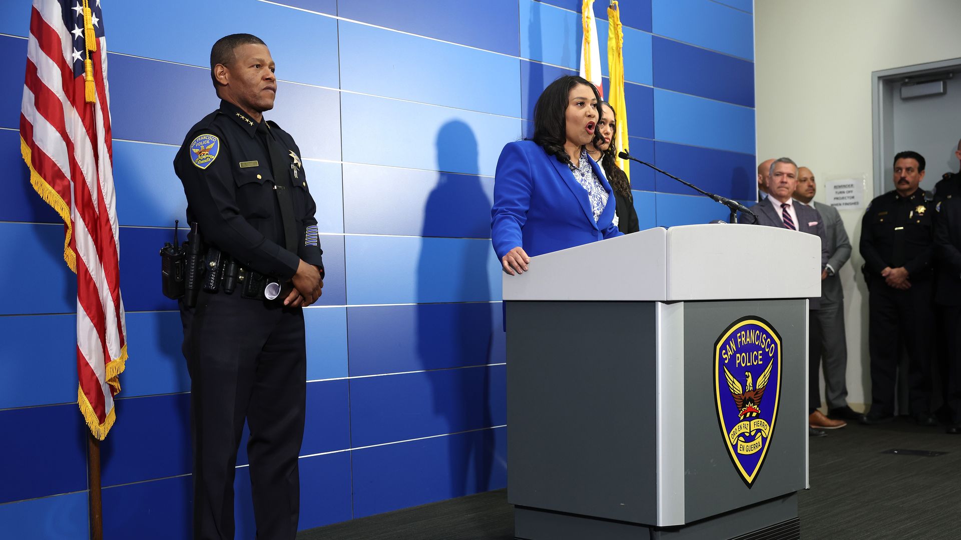Photo of London Breed speaking from a podium, surrounded by police officers, at a press conference