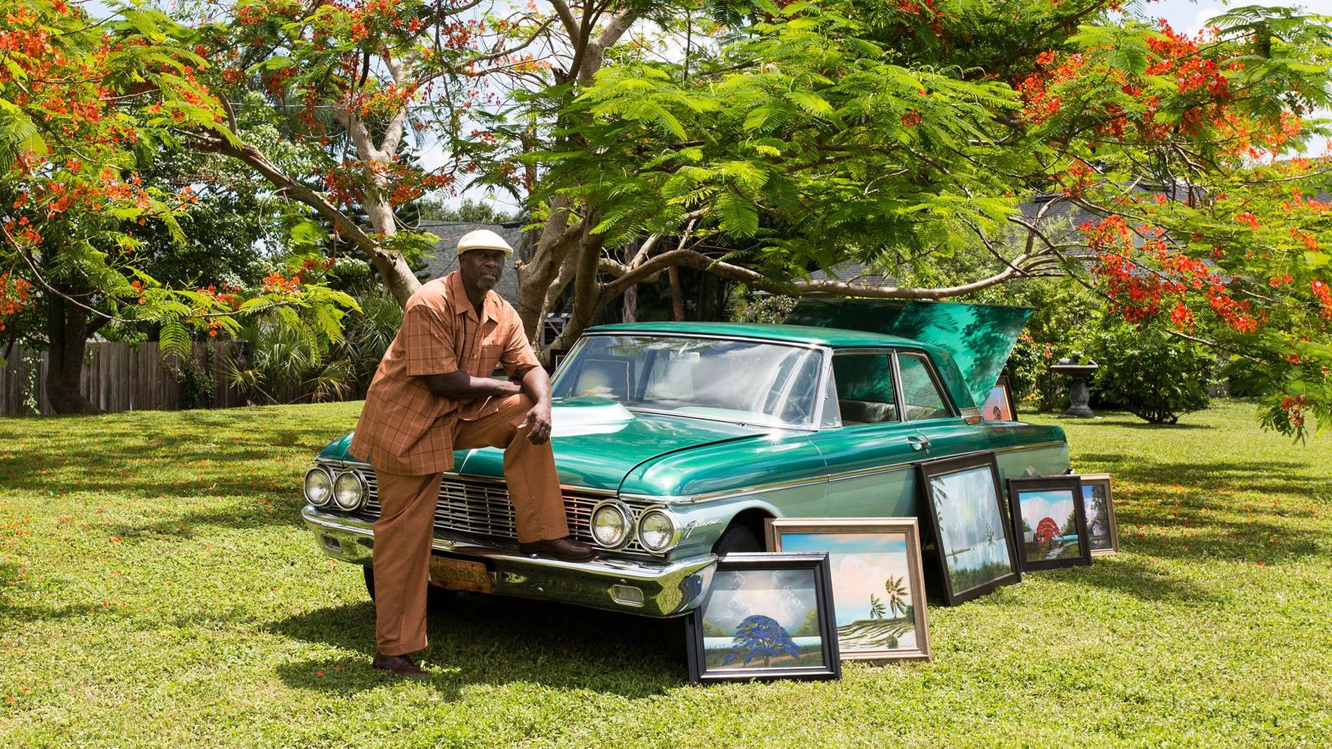 A man in a brow button down, pants and newspaper boy cap leaning on a blue-green vintage car. Several framed paintings of Florida landscapes lean against the car. A royal poinciana tree blooms red flowers in the background.