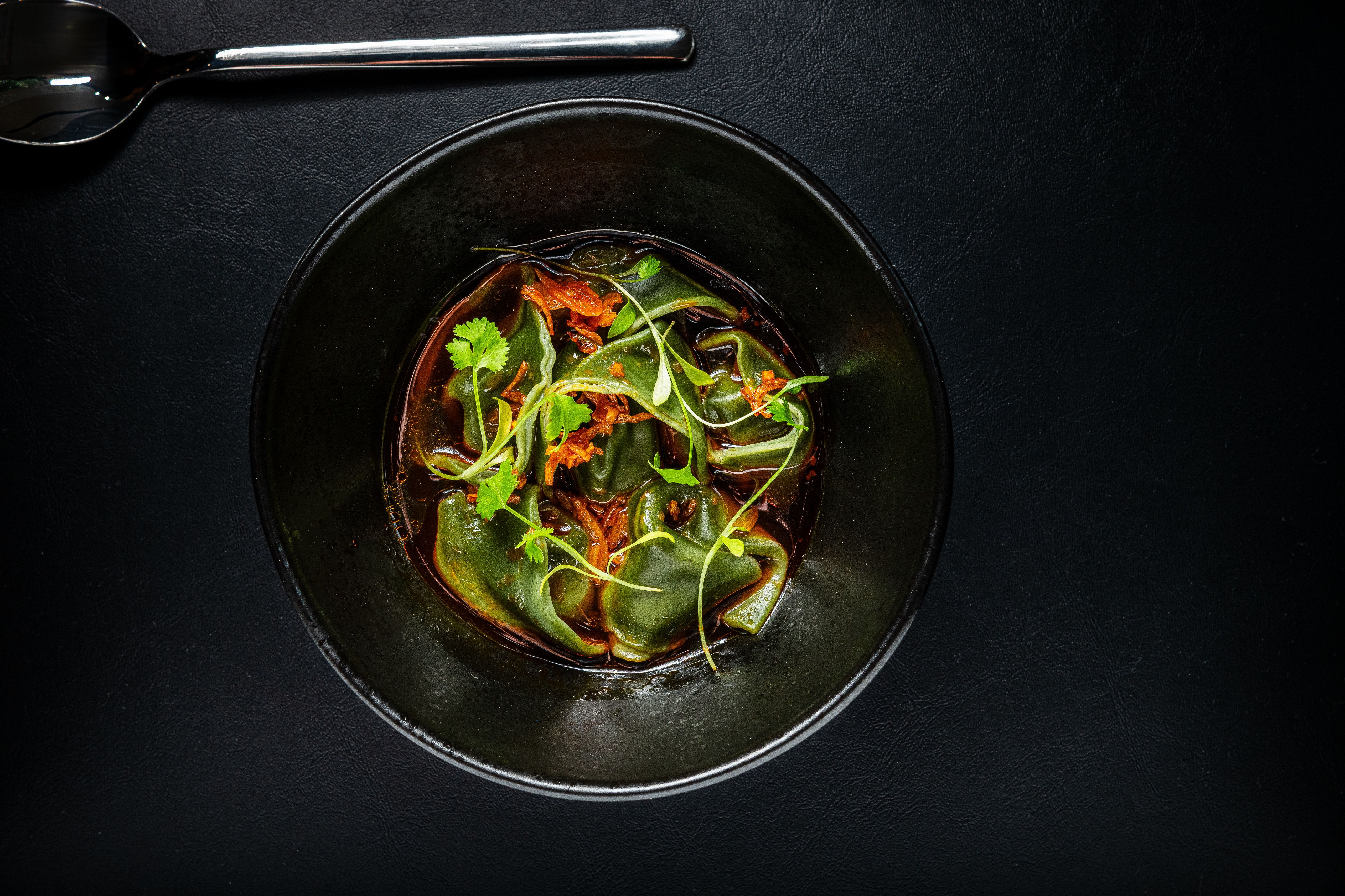 Black bowl with green dumplings in dark broth, garnished with fresh herbs and fried shallots, on a black surface with metal spoon above bowl.