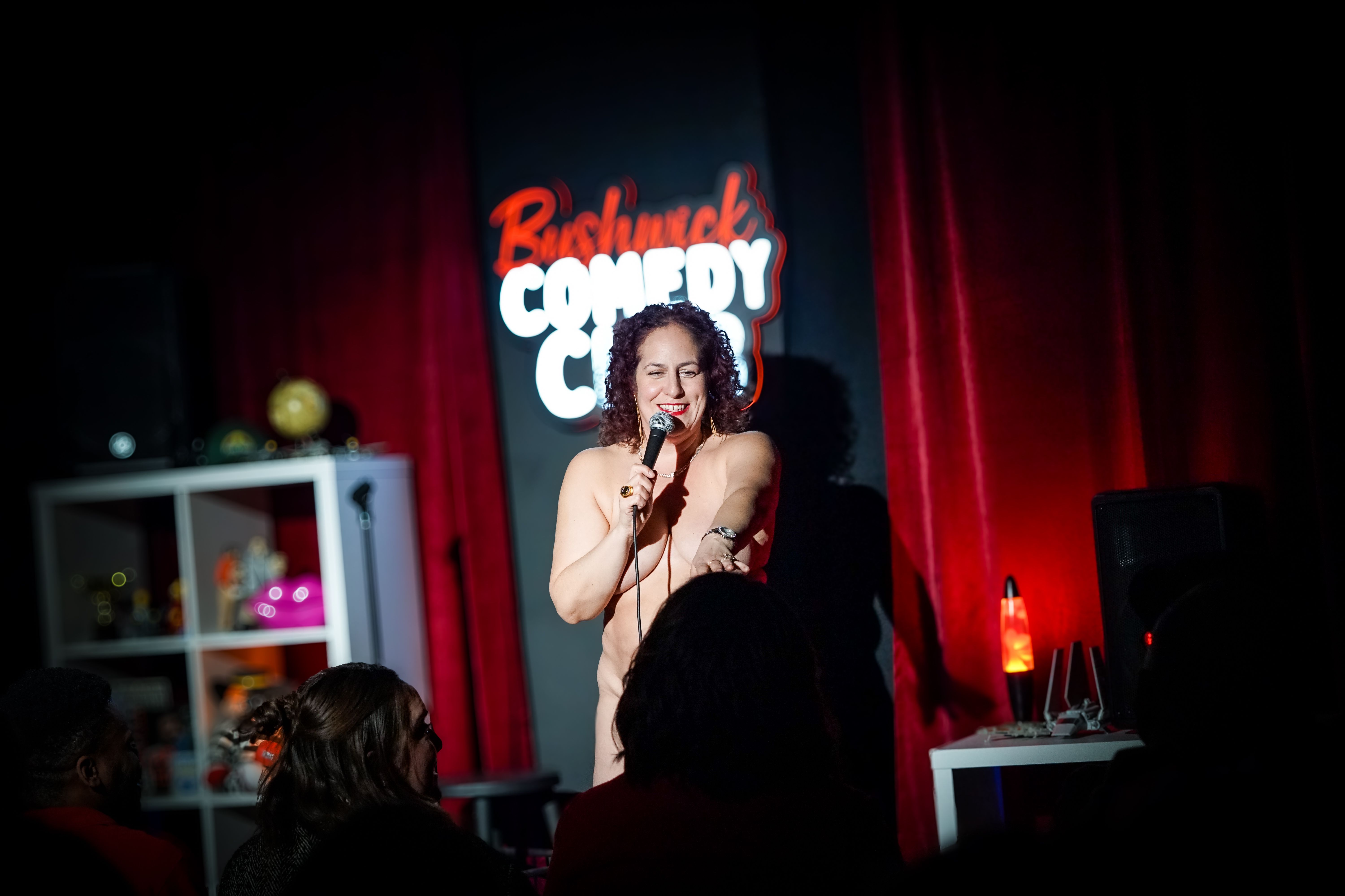 Female stand-up comedian on a small stage with a microphone, smiling. Behind her, a neon sign says "British Comedy"; red curtains frame the dark venue, with audience silhouettes in the foreground.