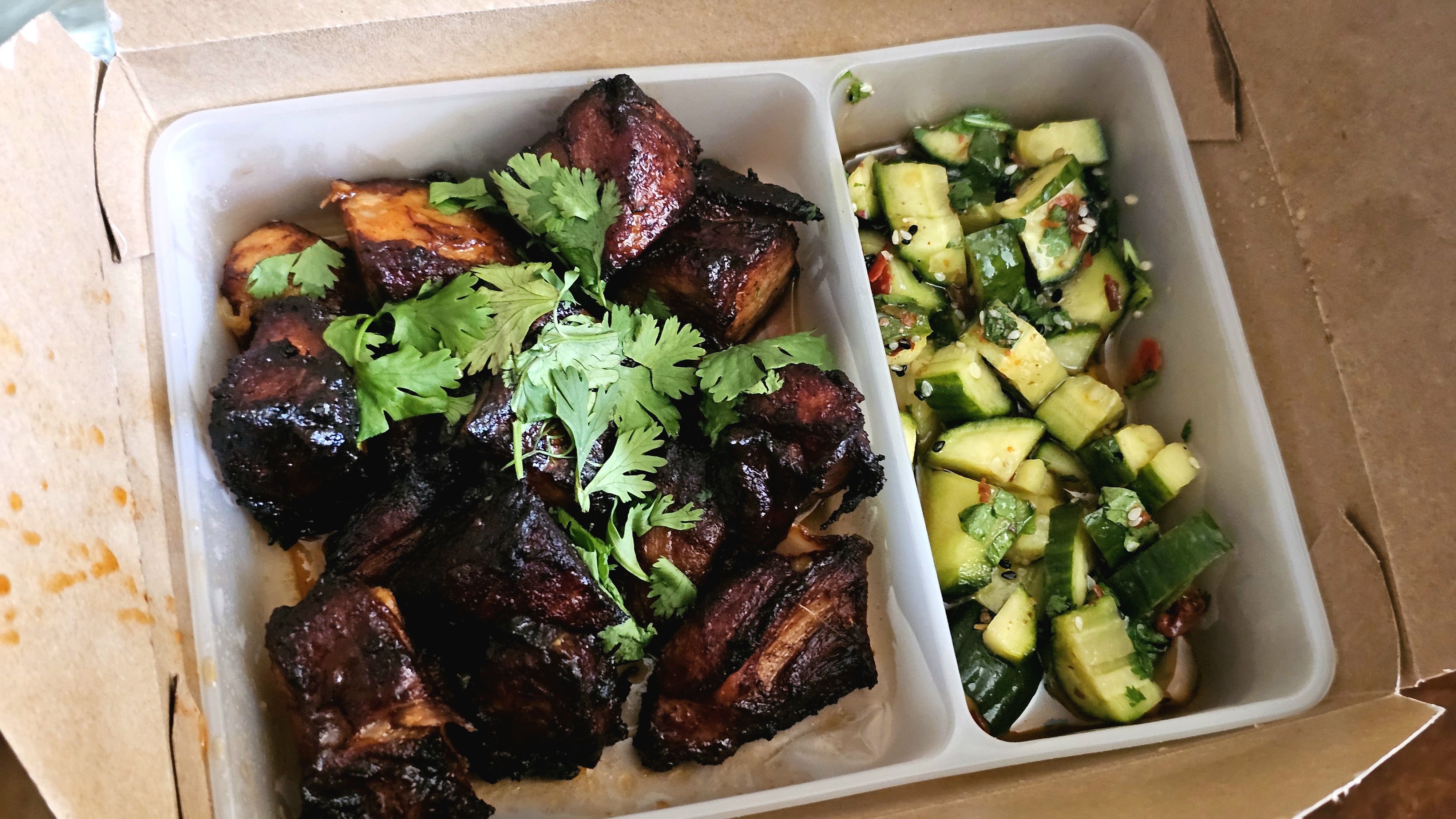 Takeout box with two compartments: left shows glossy, charred pork ribs topped with cilantro; right is a cucumber salad with sesame seeds and chopped herbs.