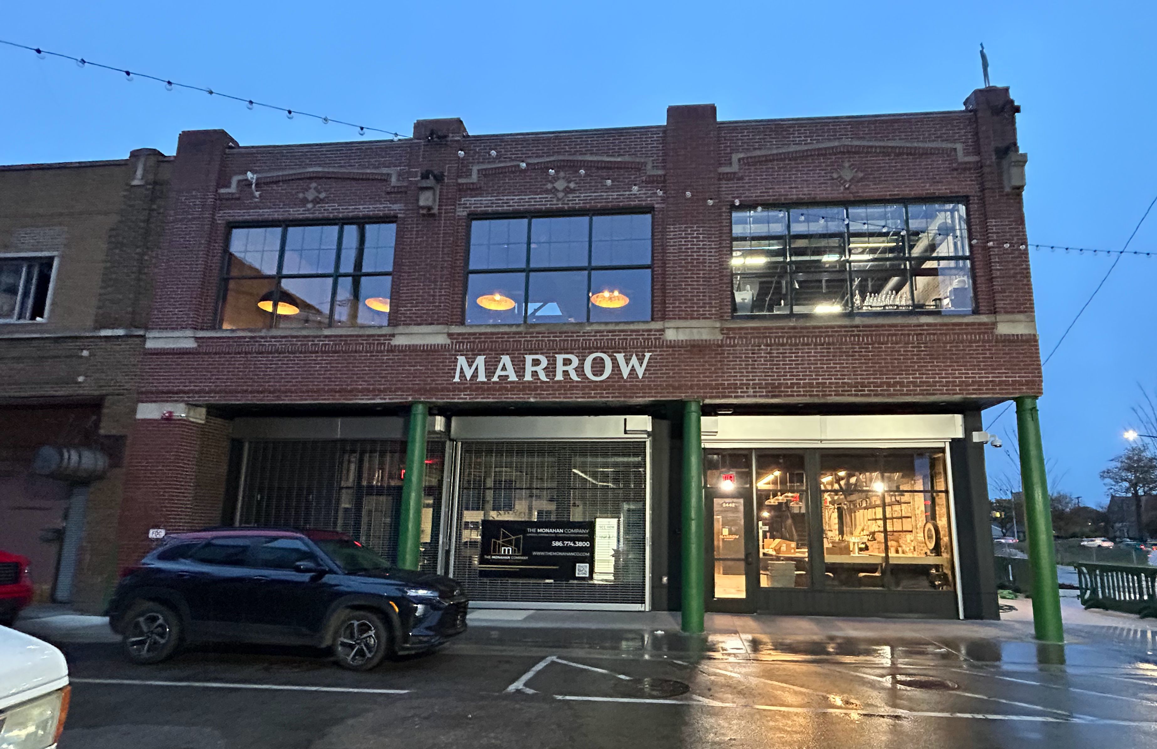 Evening view of a two-story red brick building with green columns and large windows showing lit interior, with the word MARROW displayed above the entrance.