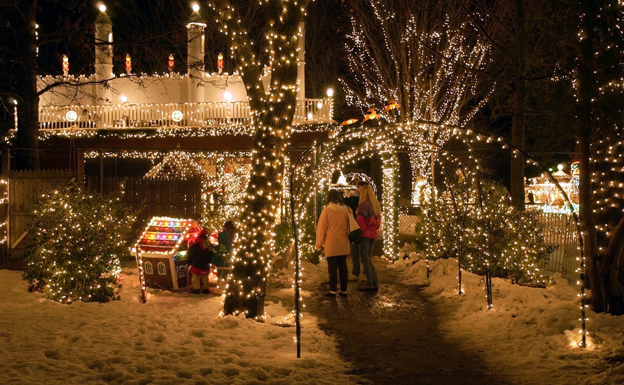 Several structures and trees light up in Christmas lights, and a couple people walking on a shoveled path.