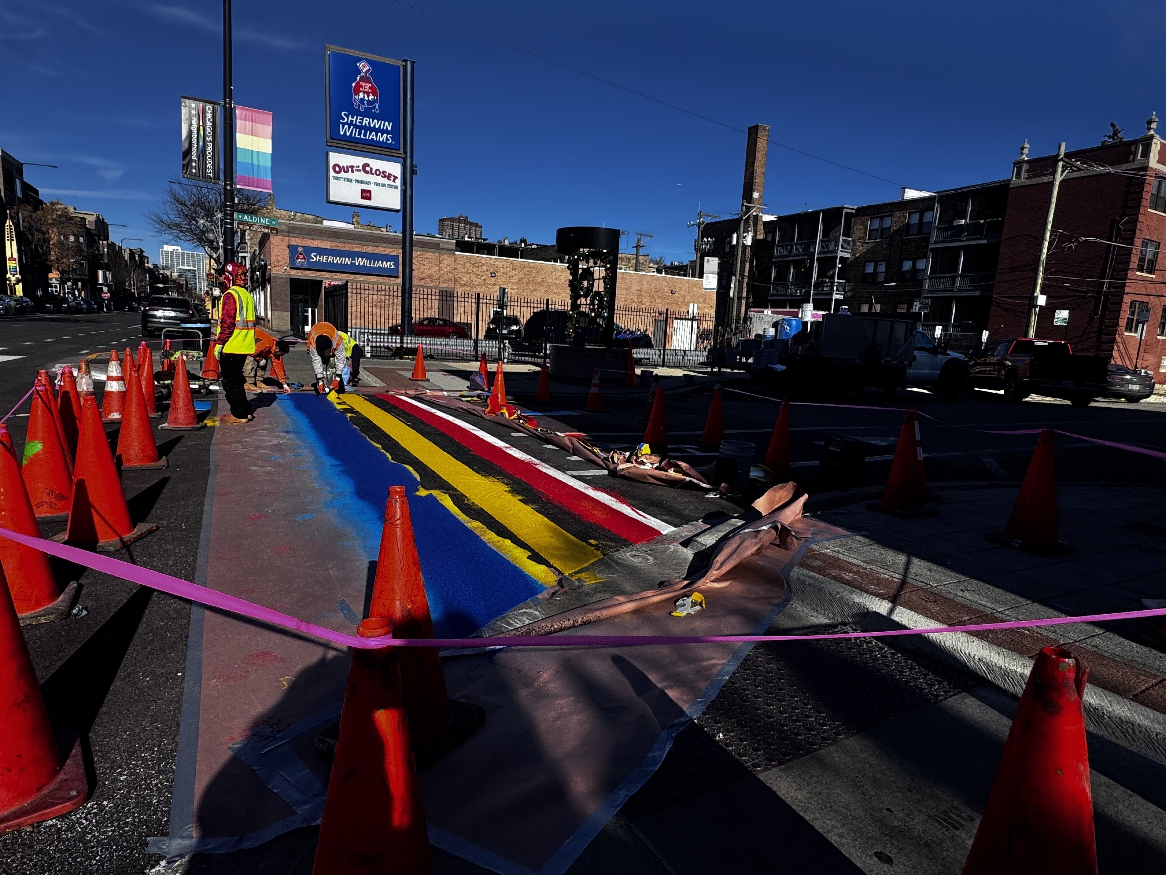 Workers in orange safety vests paint a multicolored mural on a street section, surrounded by orange cones and pink caution tape, near a Sherwin-Williams store on a clear blue day.