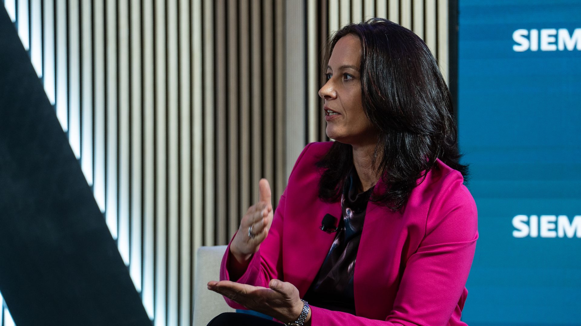 Side profile of a woman with dark hair wearing a bright pink blazer, speaking in an interview with a lapel mic, gesturing with her hands. A blue Siemens backdrop is visible behind her.