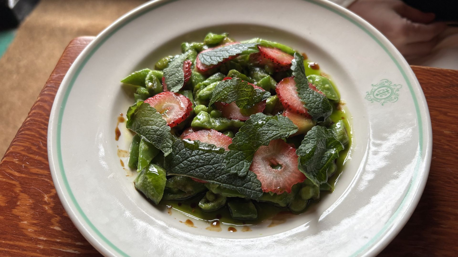 A white plate with a green rim sits on a wooden table, topped with a fresh salad of chopped green vegetables, mint leaves, and thinly sliced strawberries with a light dressing.