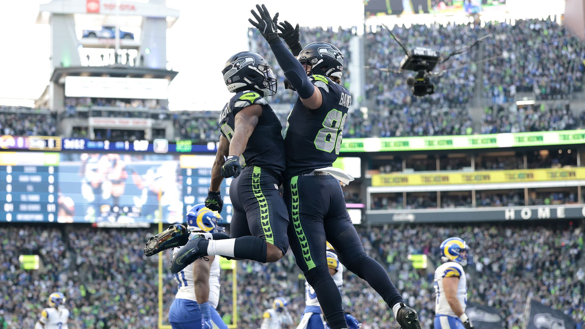 Two Seahawks players in navy and green uniforms leap into the air and bump chests in celebration on the field during a game against the Rams.