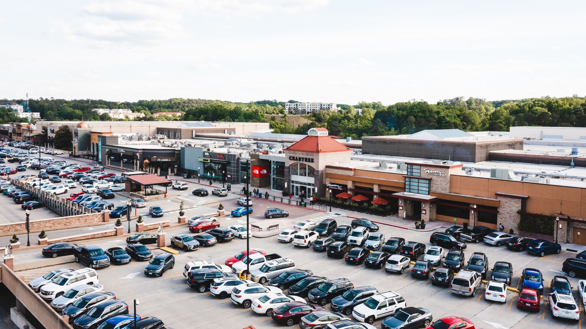 A large parking lot filled with various cars in front of a shopping center with stores like The North Face, Cracker Barrel, and other shops, surrounded by green trees under a partly cloudy sky.