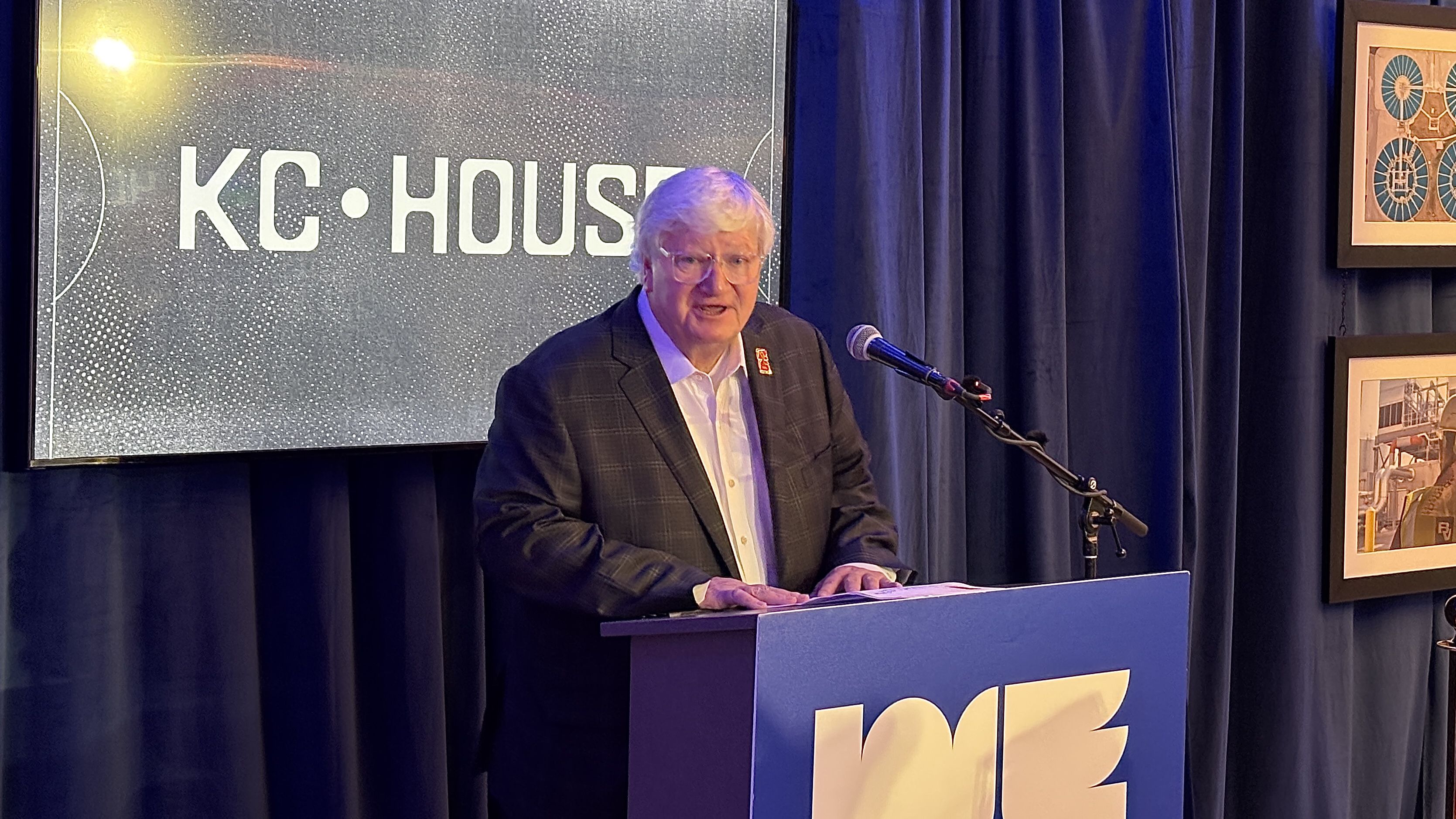Cliff Illig, a man with white hair and glasses, speaks at a blue podium with a microphone, in front of dark blue curtains and a large screen reading "KC House" with a speckled background; framed art to the right.