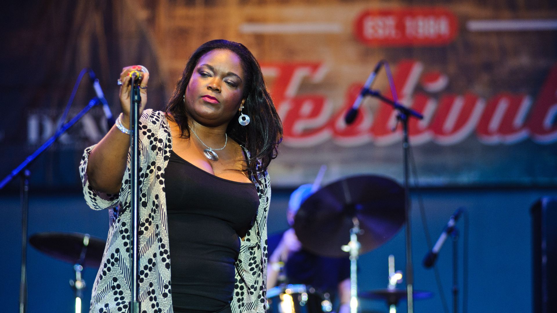 Woman on stage holding the top of a stand-up microphone. Drum set in the background.