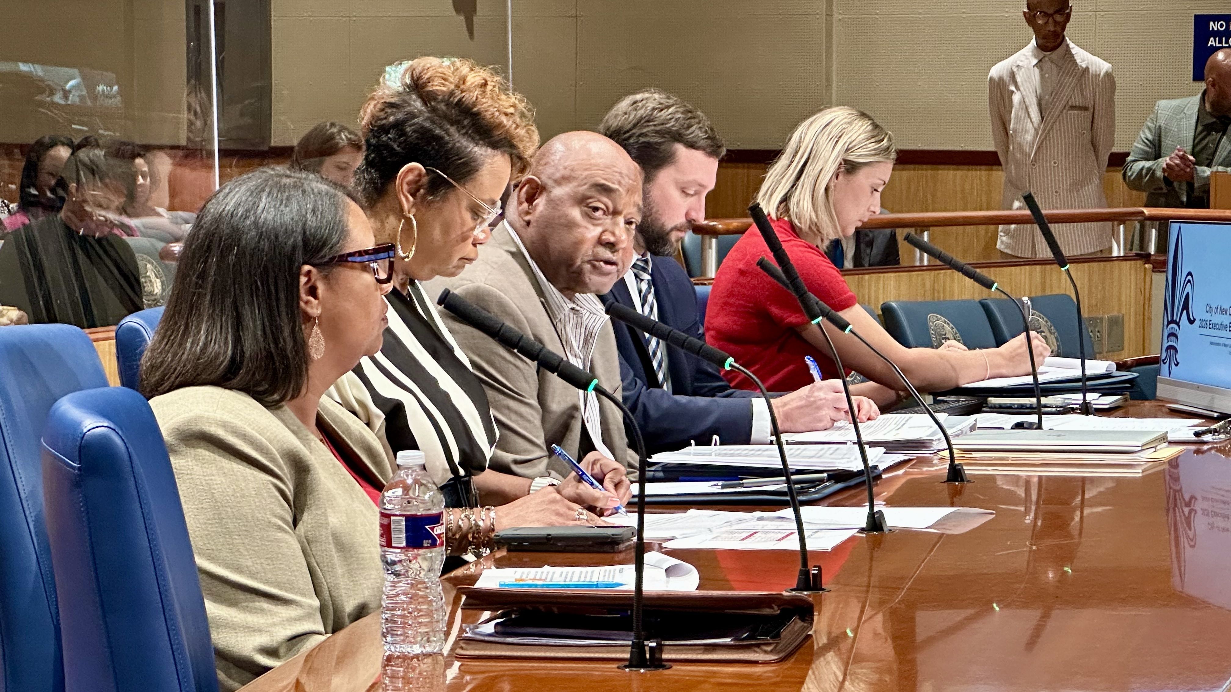 Five people seated at a conference table with microphones, taking notes and speaking in a formal meeting room with wood paneling and blue chairs.