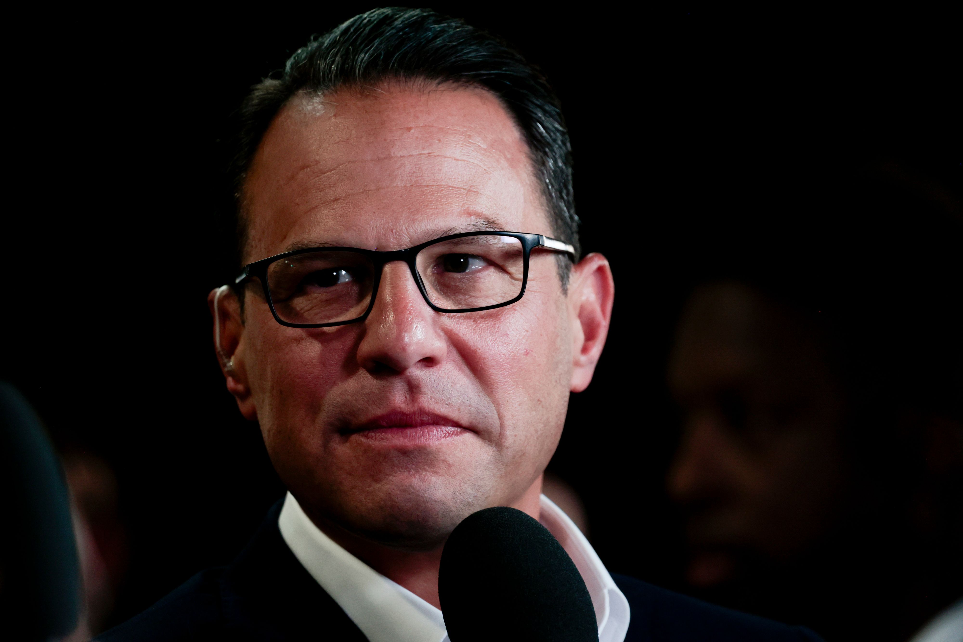 Close-up of a man with dark hair and black-framed glasses, speaking into a microphone against a black background.