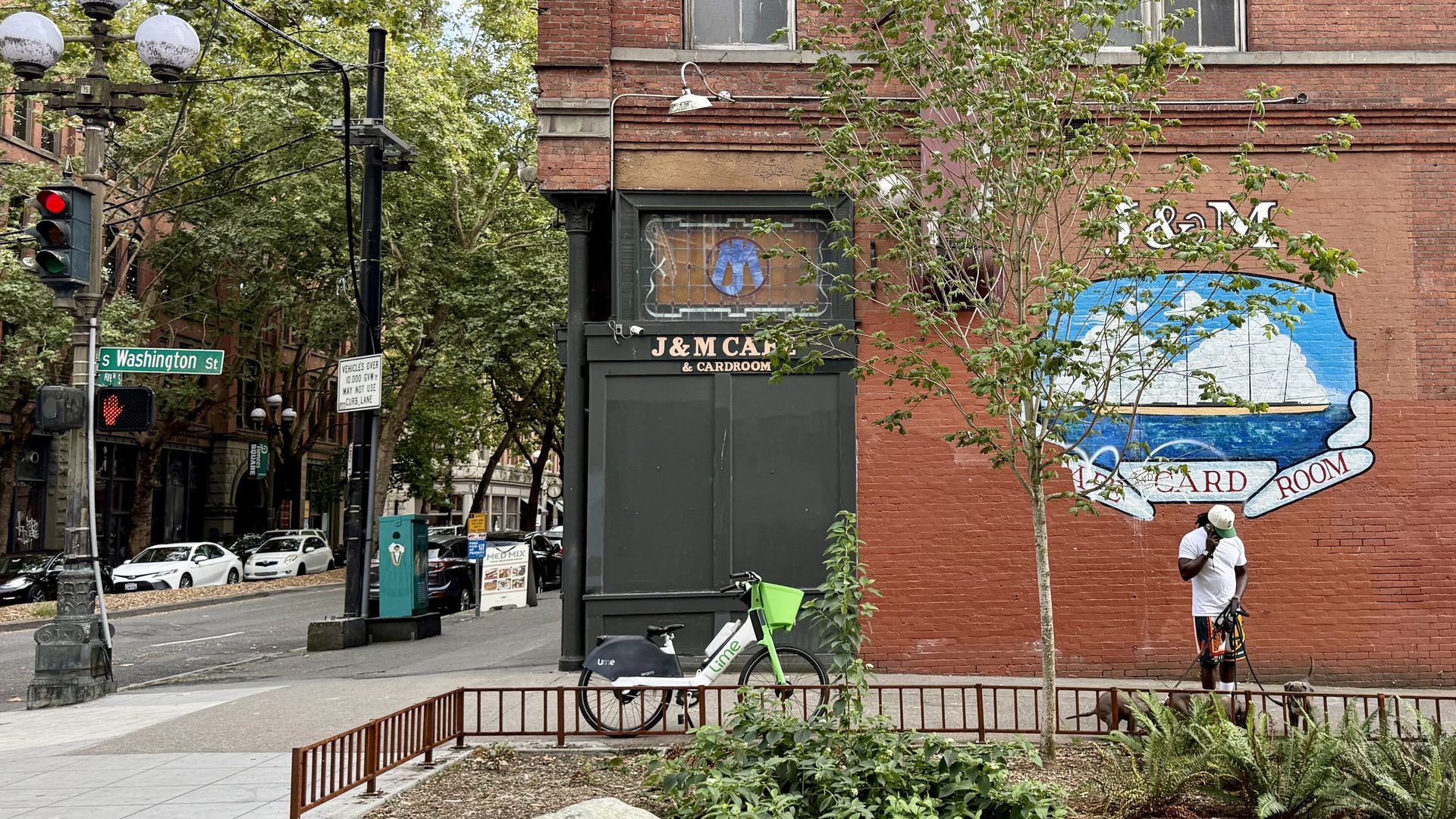 Brick building with J&M Cafe & Cardroom mural showing a ship, a man in white stands nearby, a green Lime bike is parked, street signs and a traffic light for S Washington St visible.