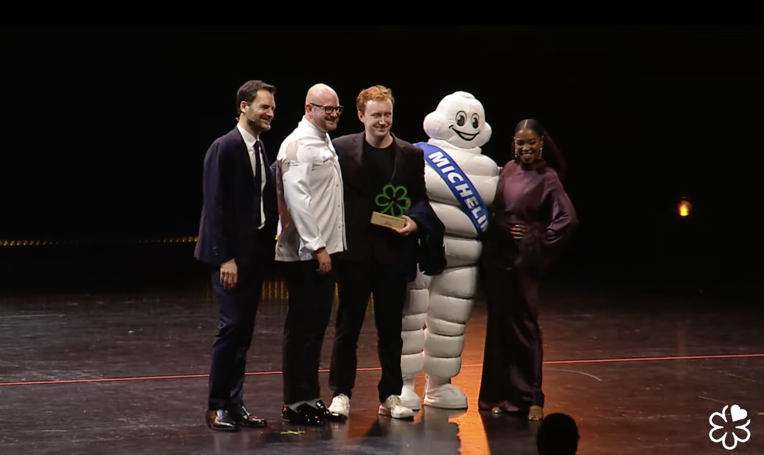 Five people stand on a stage, including a man holding a green clover-shaped award and the Michelin Man mascot wearing a blue sash reading "MICHELIN." The background is dark.
