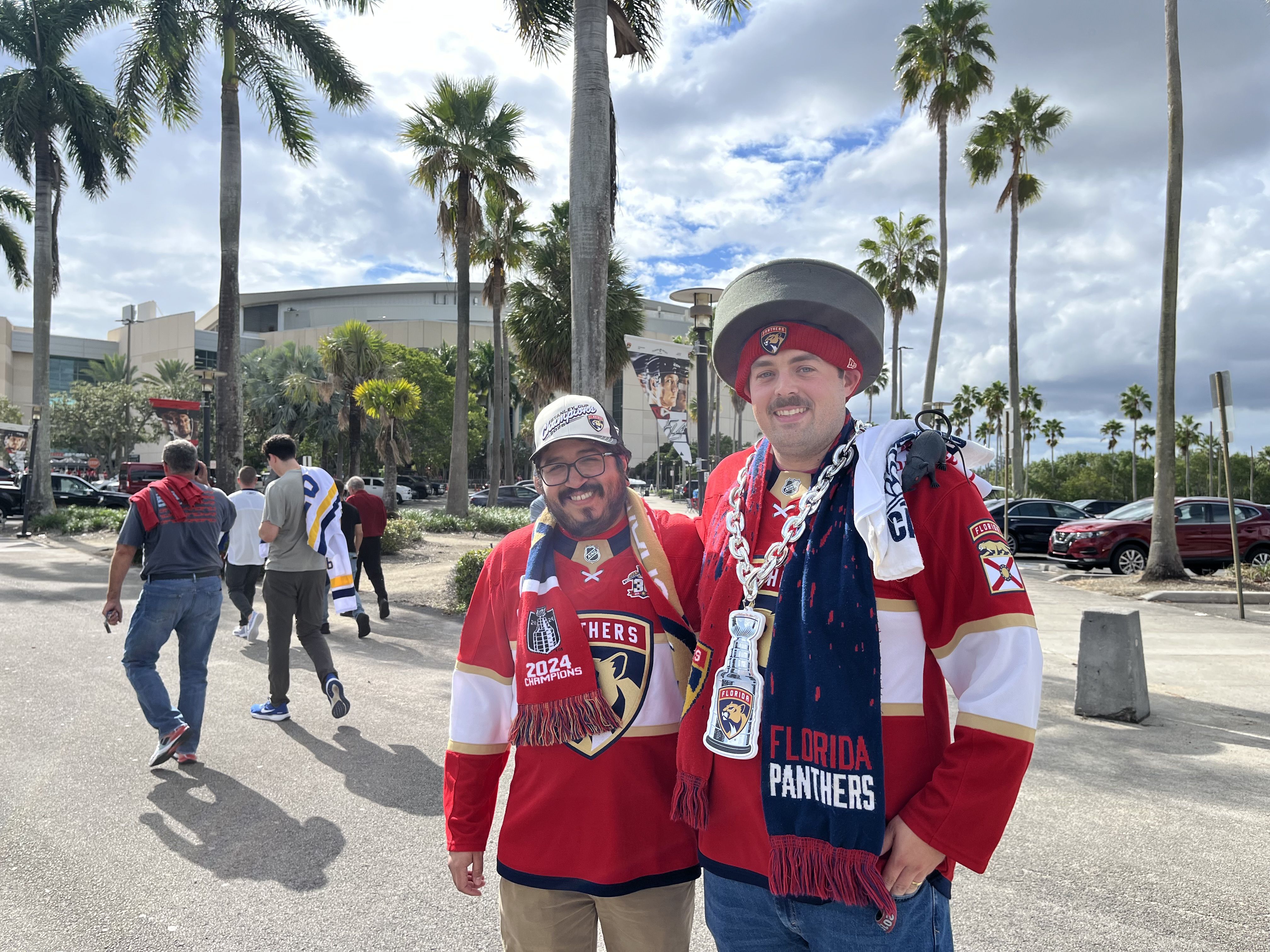 Two men wearing red Florida Panthers hockey jerseys and scarves stand smiling outdoors with palm trees and a stadium in the background on a partly cloudy day.