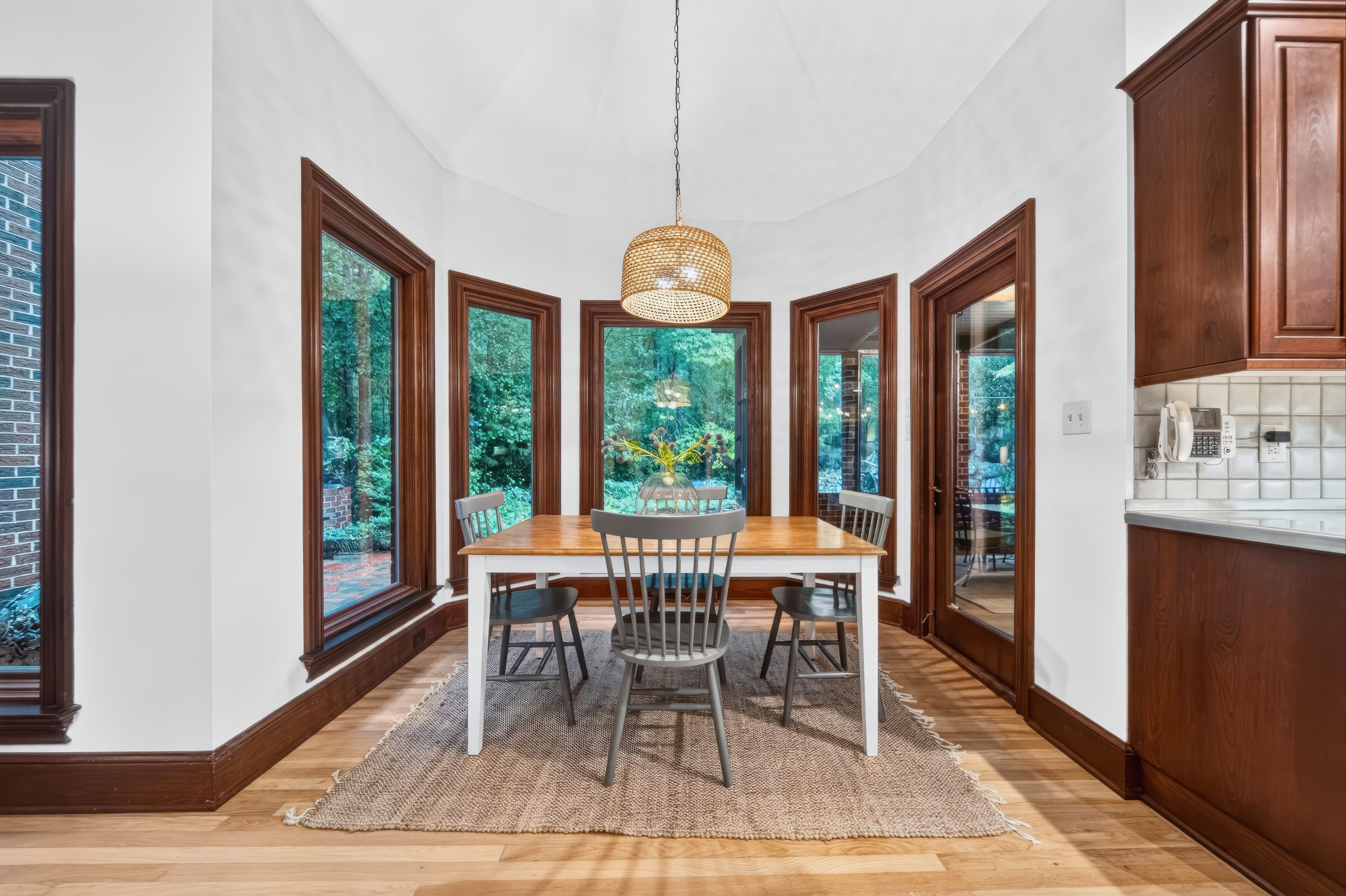 Dining nook with a wooden table and gray chairs under a woven pendant light. A potted plant sits on the table; bay windows with dark wood trim, white walls, and a rug; kitchen cabinets to the right.
