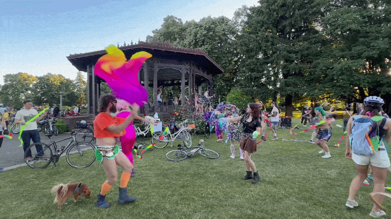 People dance and twirl colorful ribbons on the lawn around the Peninsula Park bandstand during a summer bike-themed gathering. Bicycles line the grass as attendees in bright outfits celebrate in the evening light.