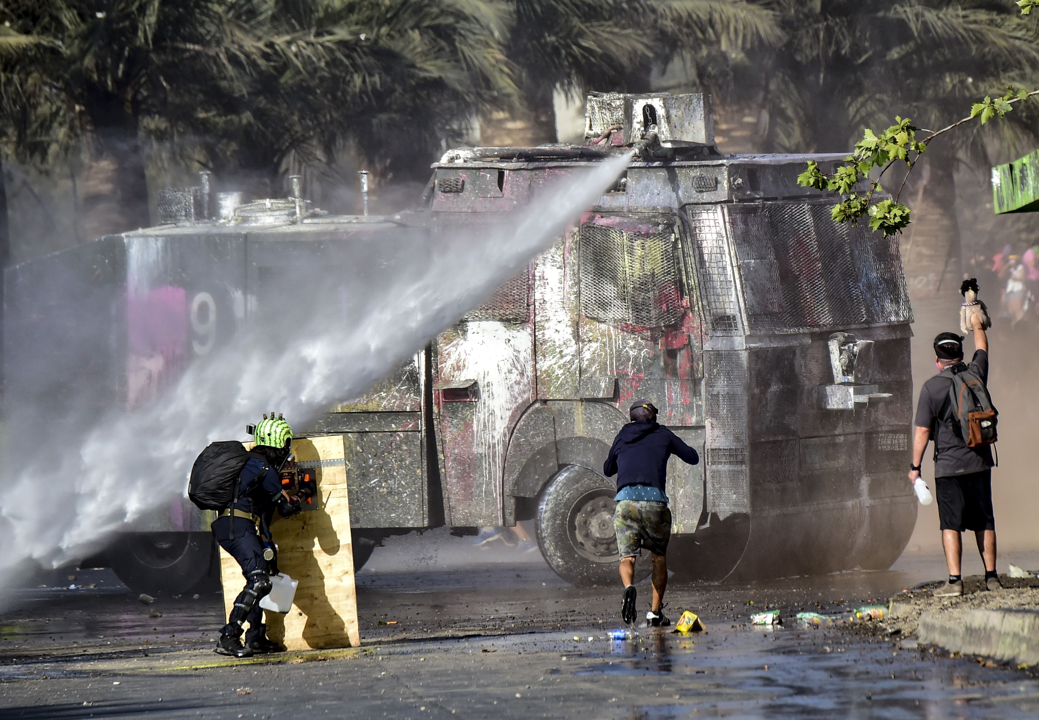 In this image, a truck sprays a water hose at a protestor who uses a plank of wood as a shield 