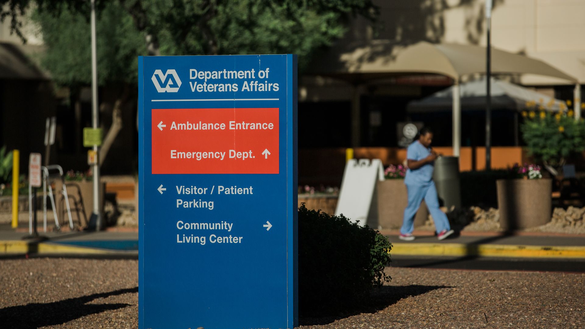 A blue and red sign in front of a VA hospital that informs people locations of different departments.