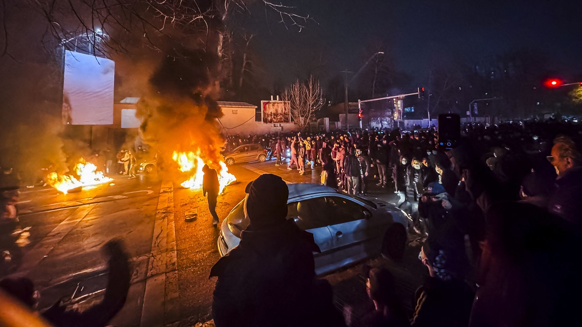 Nighttime street scene with large crowd of people gathered around burning fires and thick black smoke rising, cars nearby, and leafless trees overhead.