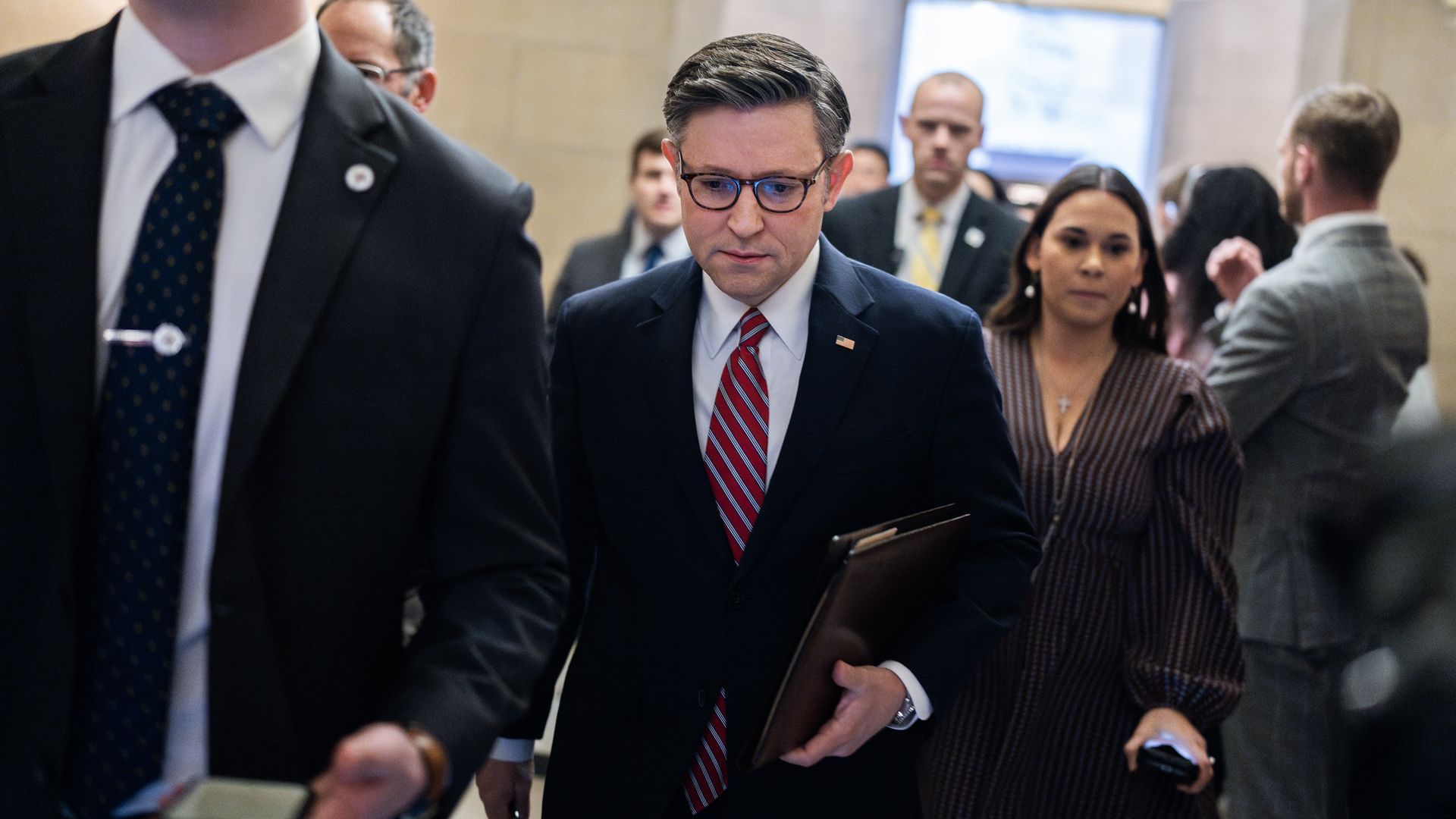 House Speaker Mike Johnson, wearing a dark blue suit, light blue shirt, red striped tie and glasses, walks with aides and his security detail.