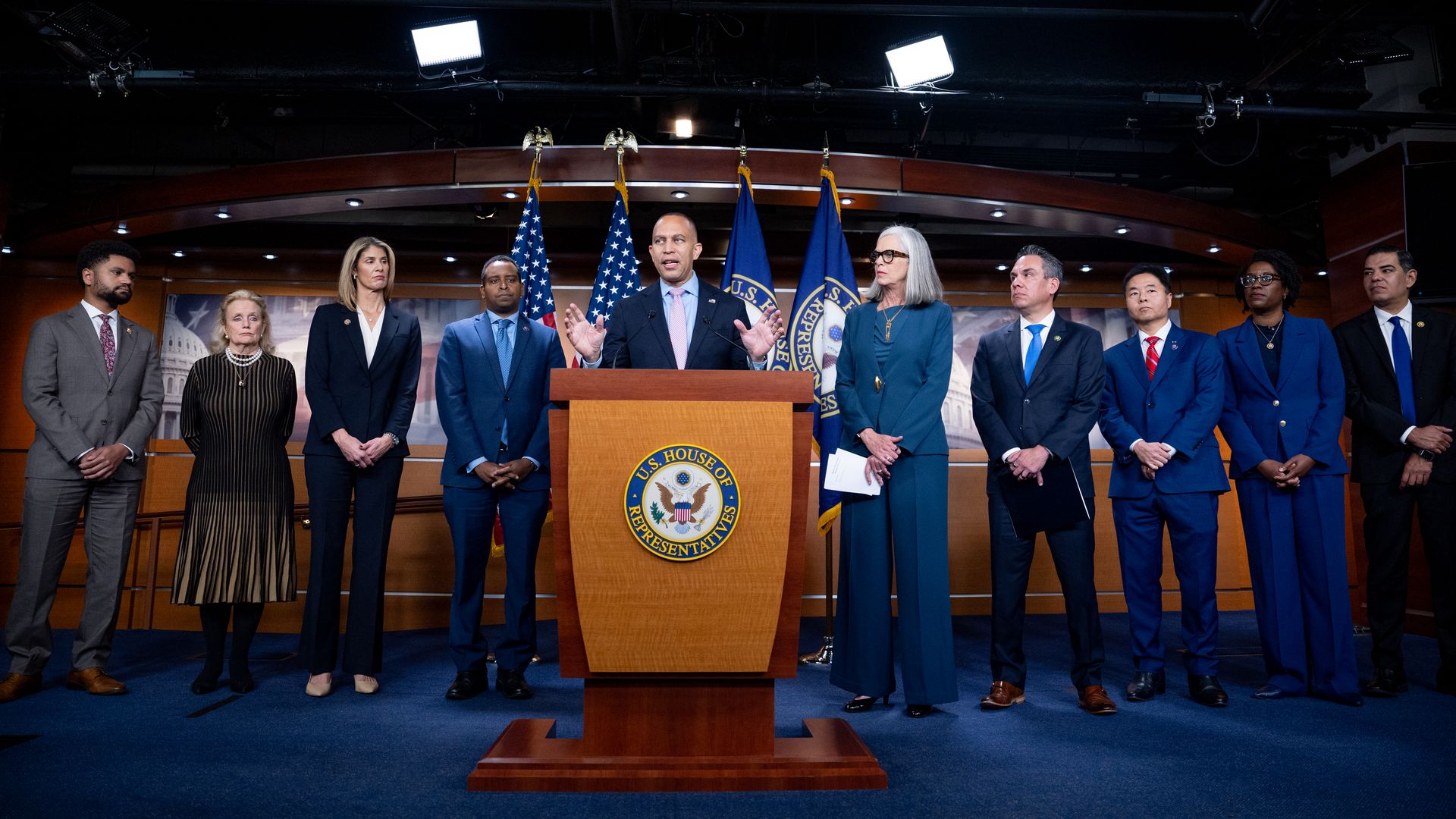 House Democratic leadership standing on a blue carpet behind a wooden podium in front of a row of flags.