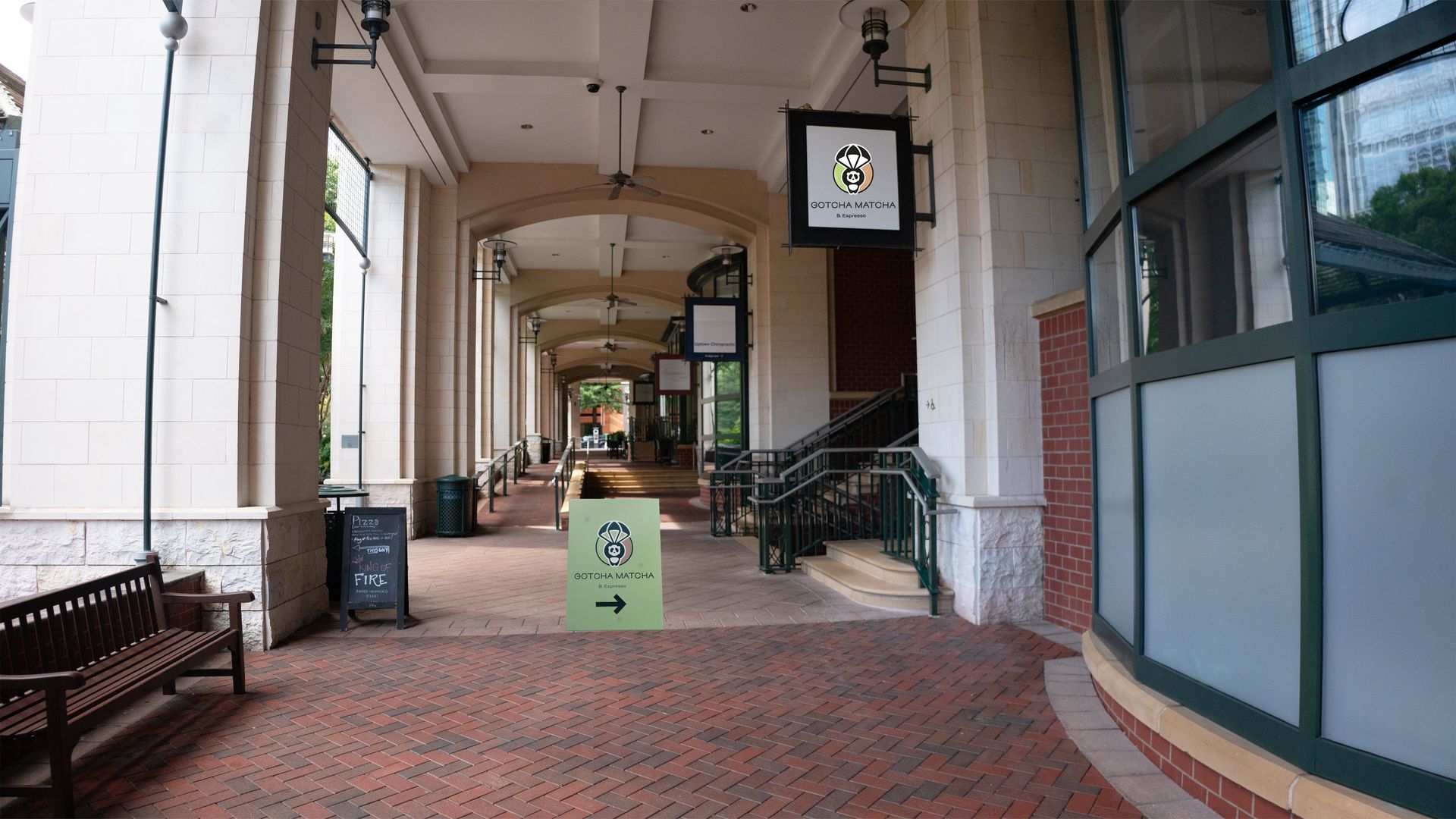 Covered outdoor corridor with brick flooring, beige columns, wooden bench, and signs for Gotcha Matcha with a panda logo, an arrow points to the right into the corridor.