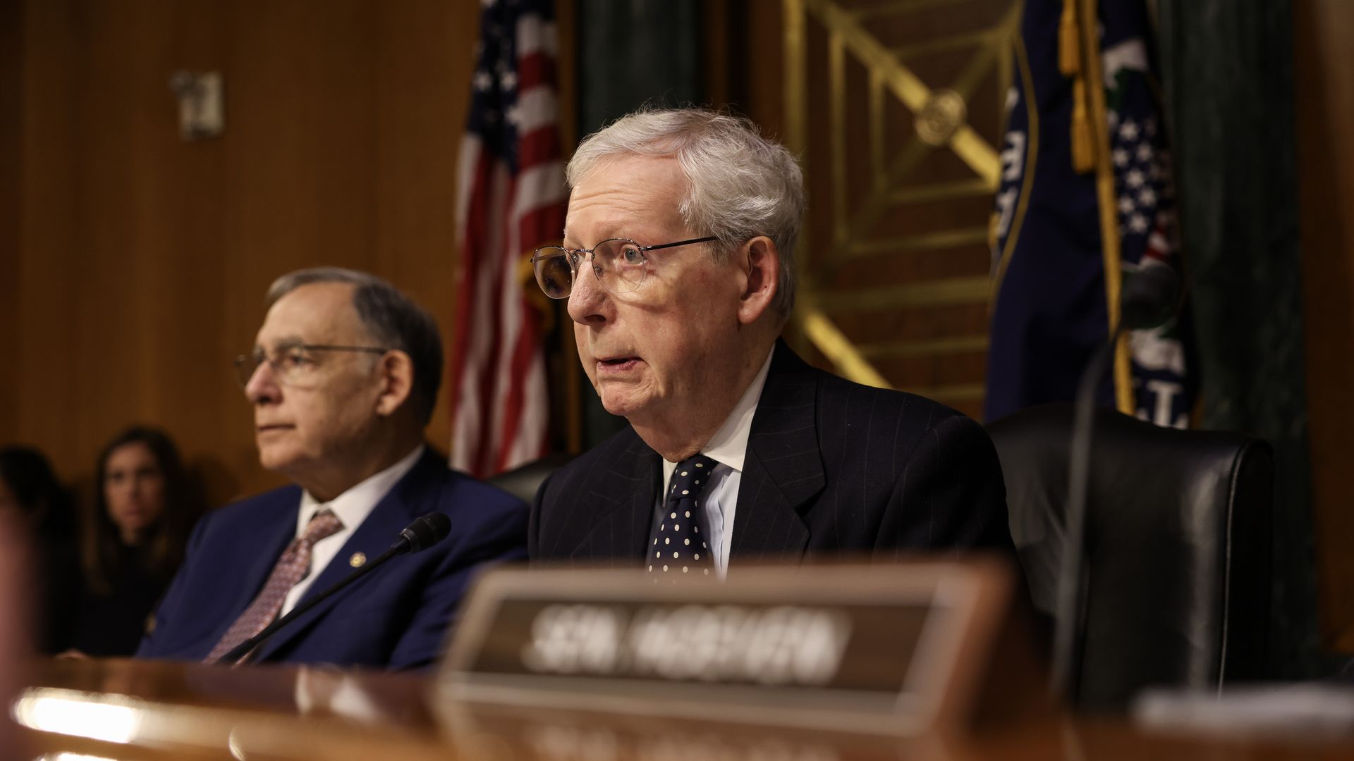 Sen. Mitch McConnell, wearing a gray suit and sitting at a committee dais speaking into a microphone in front of flags and a brown wooden wall.