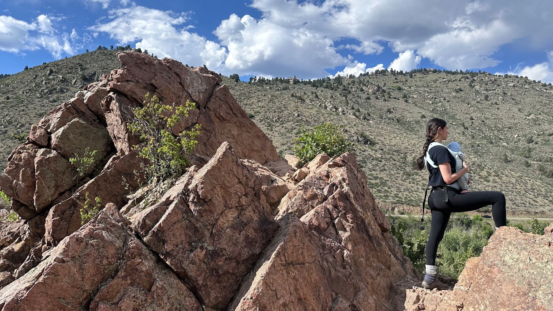 Foreground: jagged reddish rocks with a woman carrying a baby in a front carrier, standing on a rocky hillside; blue sky, white clouds, and sparse desert vegetation behind.