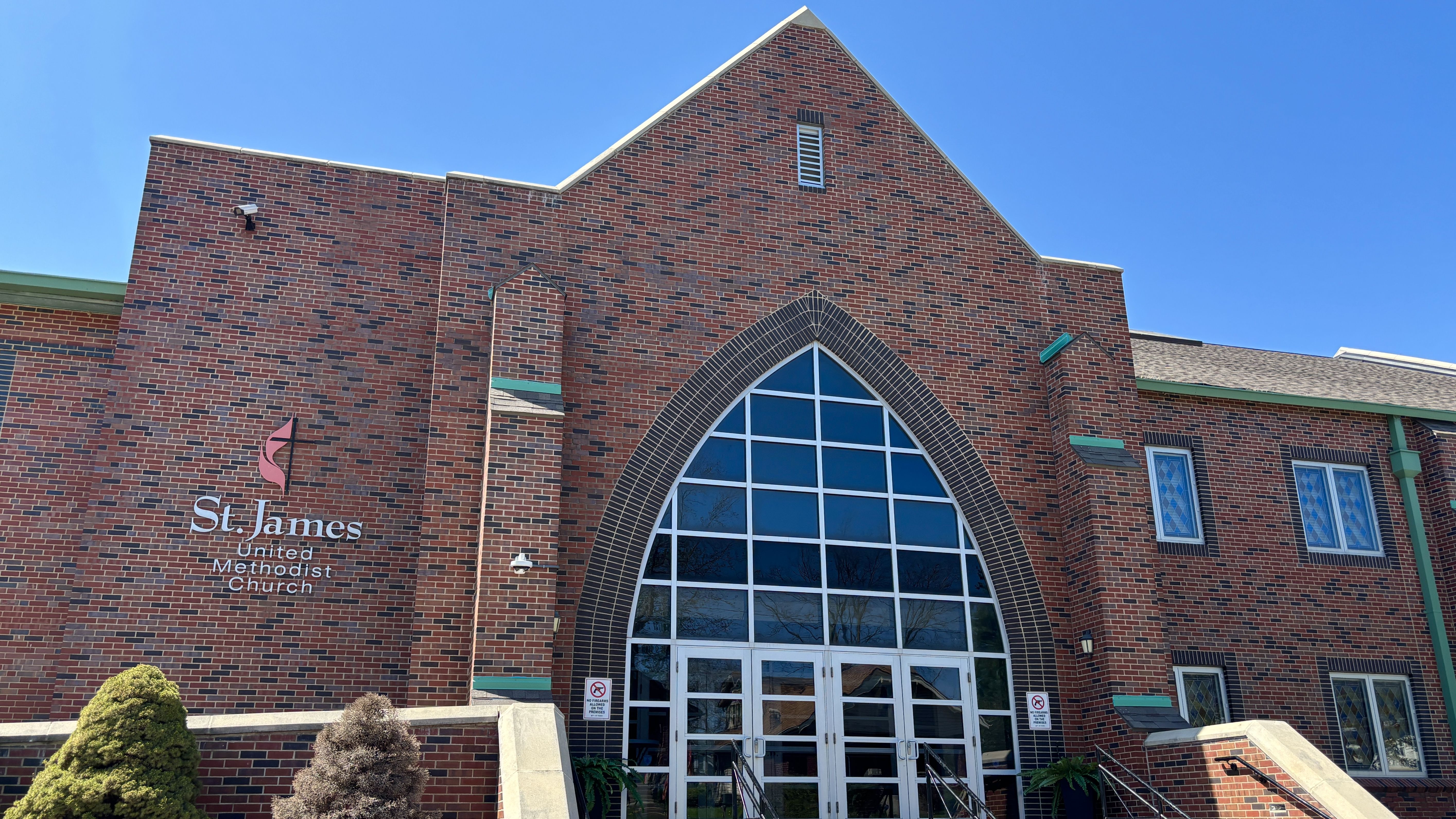 Front view of St. James United Methodist Church: red brick building with a tall pointed arch glass entrance, green trim, and a bright blue sky.
