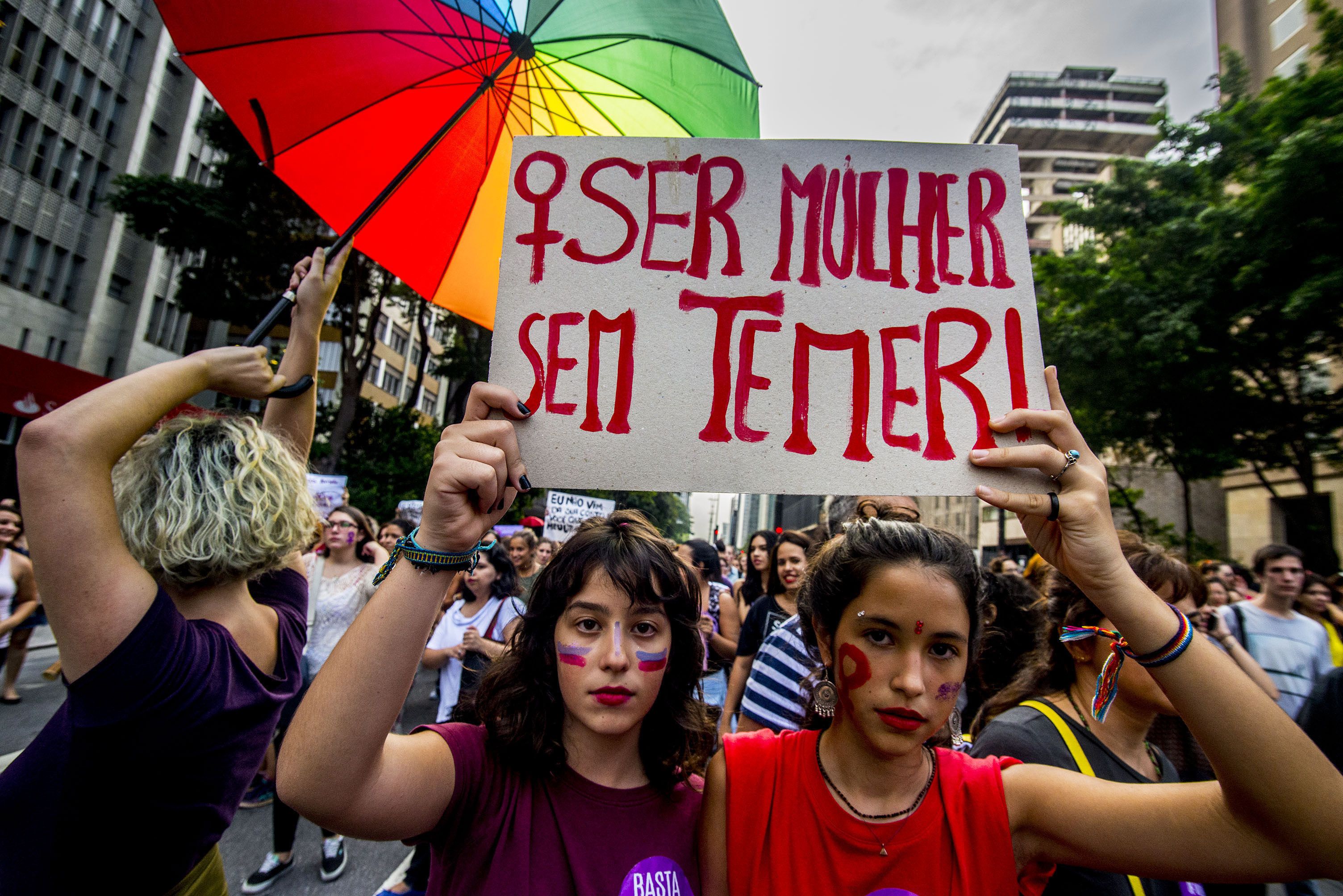 Two women march side by side wearing red and holding a sign in Portuguese that says "be a woman without fear"