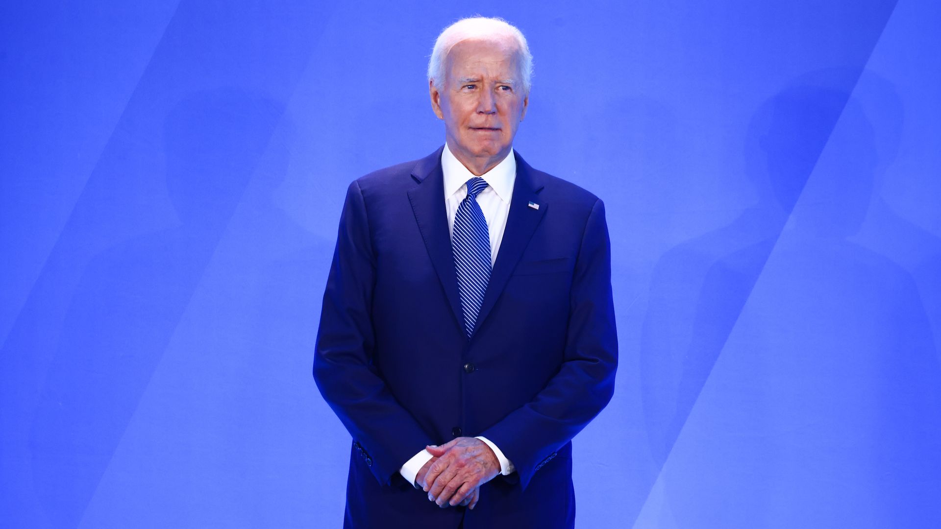 President Joe Biden, wearing a blue suit and standing in front of a blue wall with his hands clasped in front of his stomach.