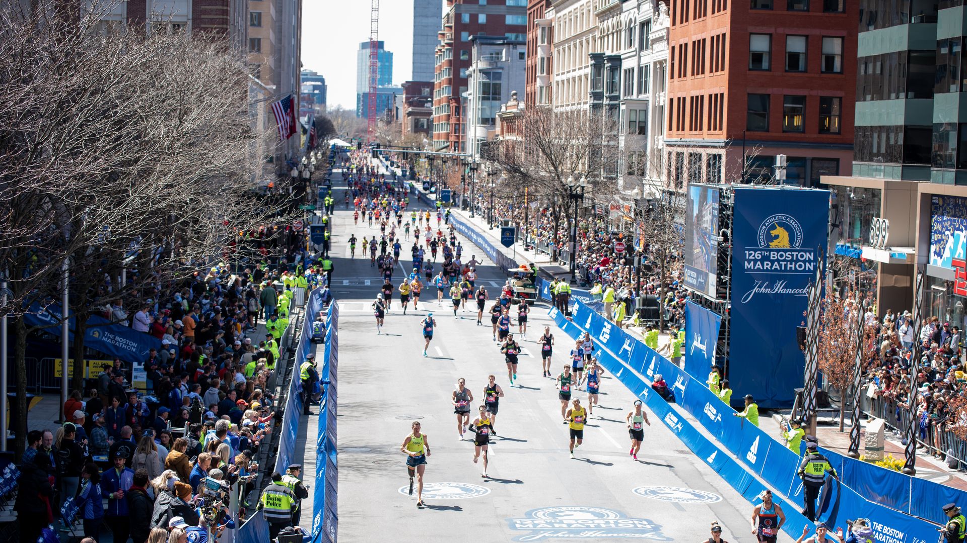 Runners on the Boston Marathon route head down the street as crowds cheer them on and watch.