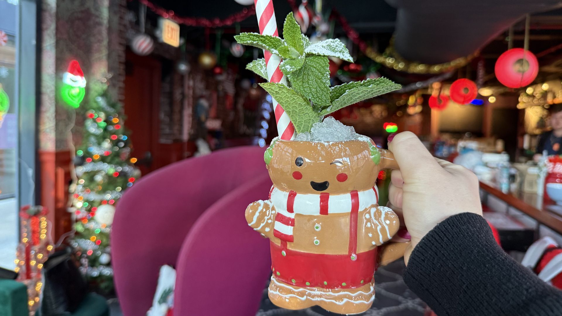 A hand holds a festive gingerbread man mug filled with a drink, crushed ice, red and white striped straw, and mint leaves, in a Christmas-decorated indoor setting with lights and ornaments.