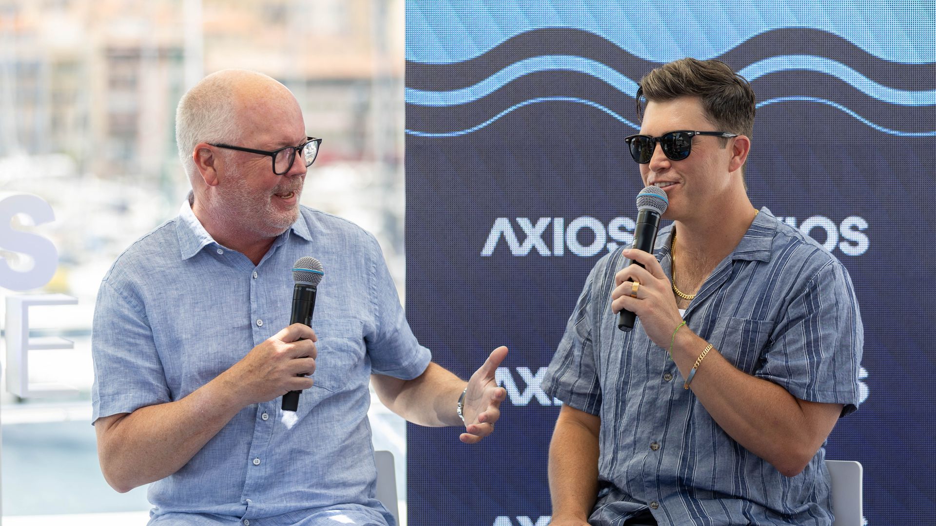 Two men on a panel speak into microphones; left man is bald with glasses wearing a light shirt, right man wears sunglasses and a striped shirt. A blue Axios banner with wave design behind them.