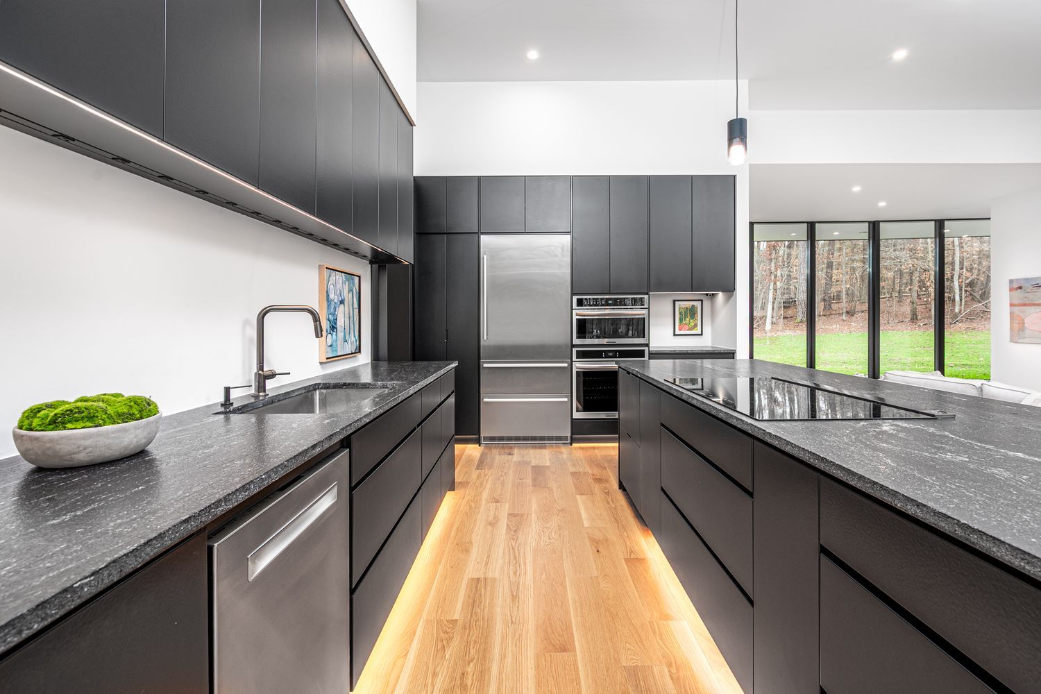 Modern kitchen with black cabinets, stainless steel appliances, black countertops, wooden floor, under-cabinet lighting, and large windows showing a green yard and trees.