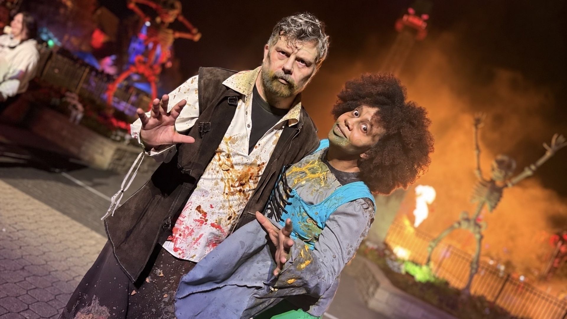 Two actors in zombie costumes pose on stage; a bearded man in a splattered white shirt and dark vest stands in front, beside a partner with an Afro wig and blue shirt, against an orange backdrop.