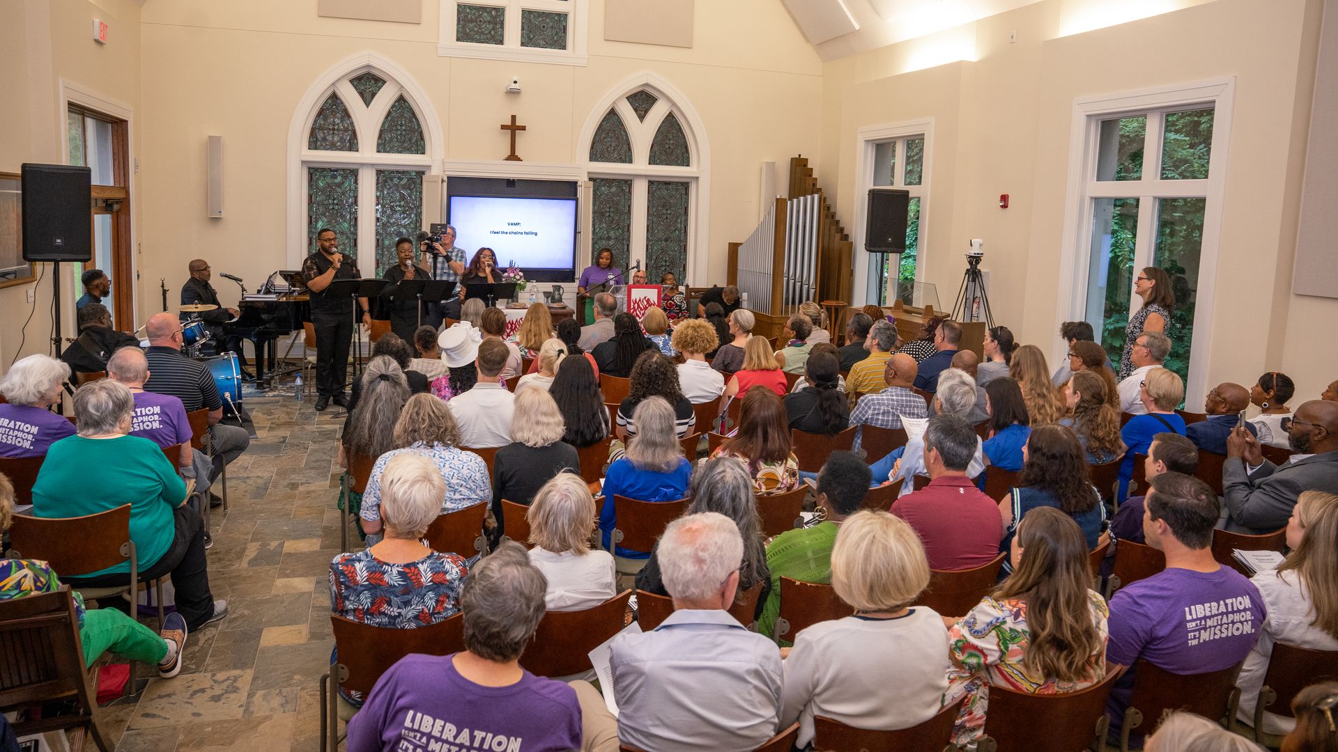 Interior of a church with a diverse audience watching musicians and singers perform at the front. Stained glass windows and a cross are visible behind performers. Some wear purple shirts.