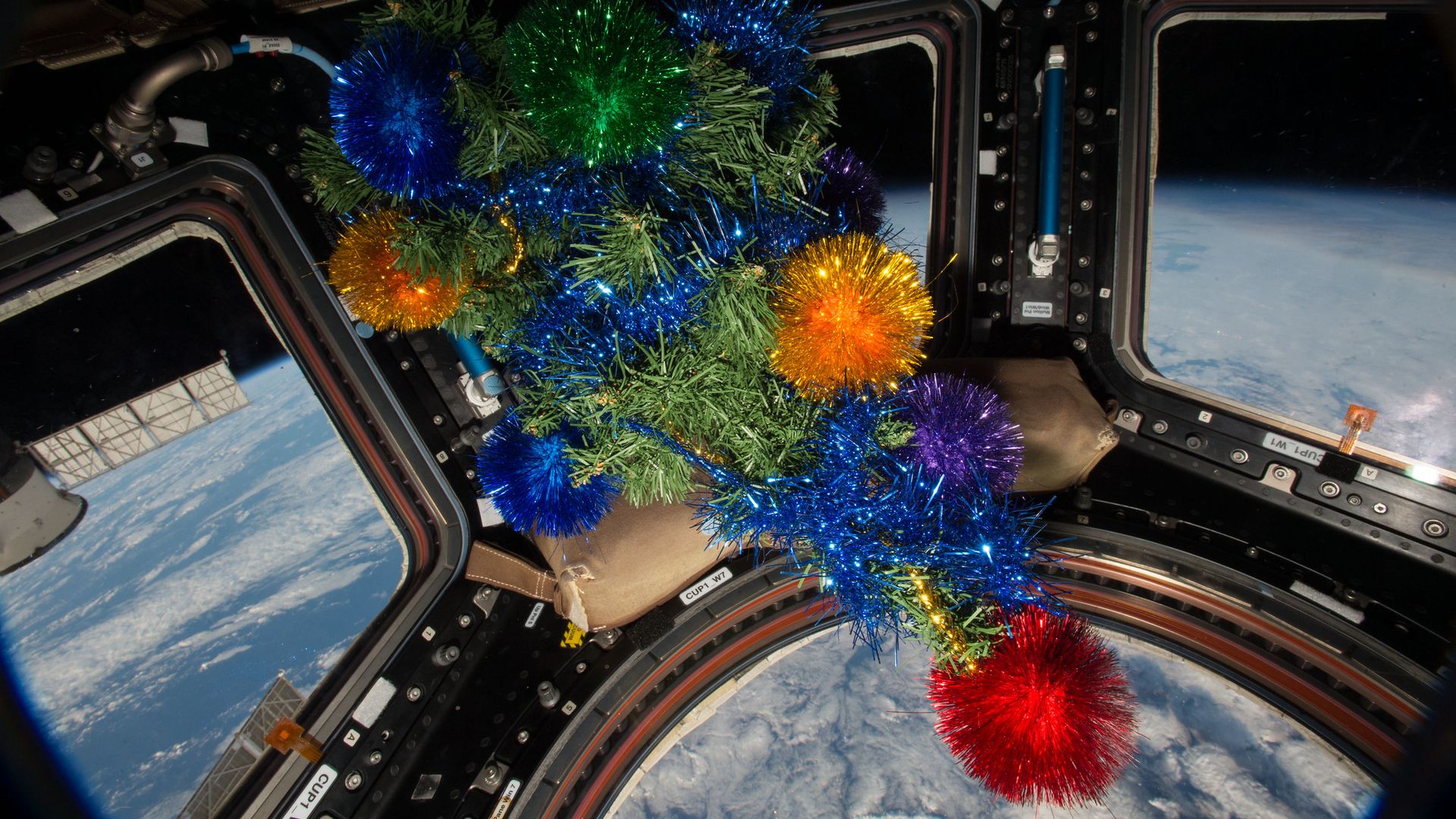 A fake Christmas tree floats in the window of the International Space Station. You can see Earth's white clouds and blue oceans under it.
