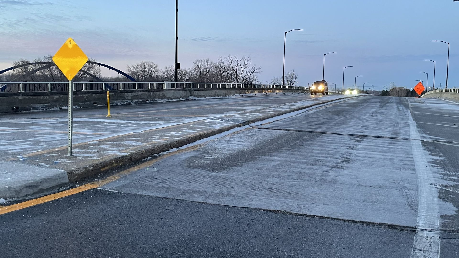 Icy winter bridge scene with a yellow diamond warning sign on the left, guard rails, leafless trees, distant cars with headlights, and an orange "Road Work Ahead" sign on the right.