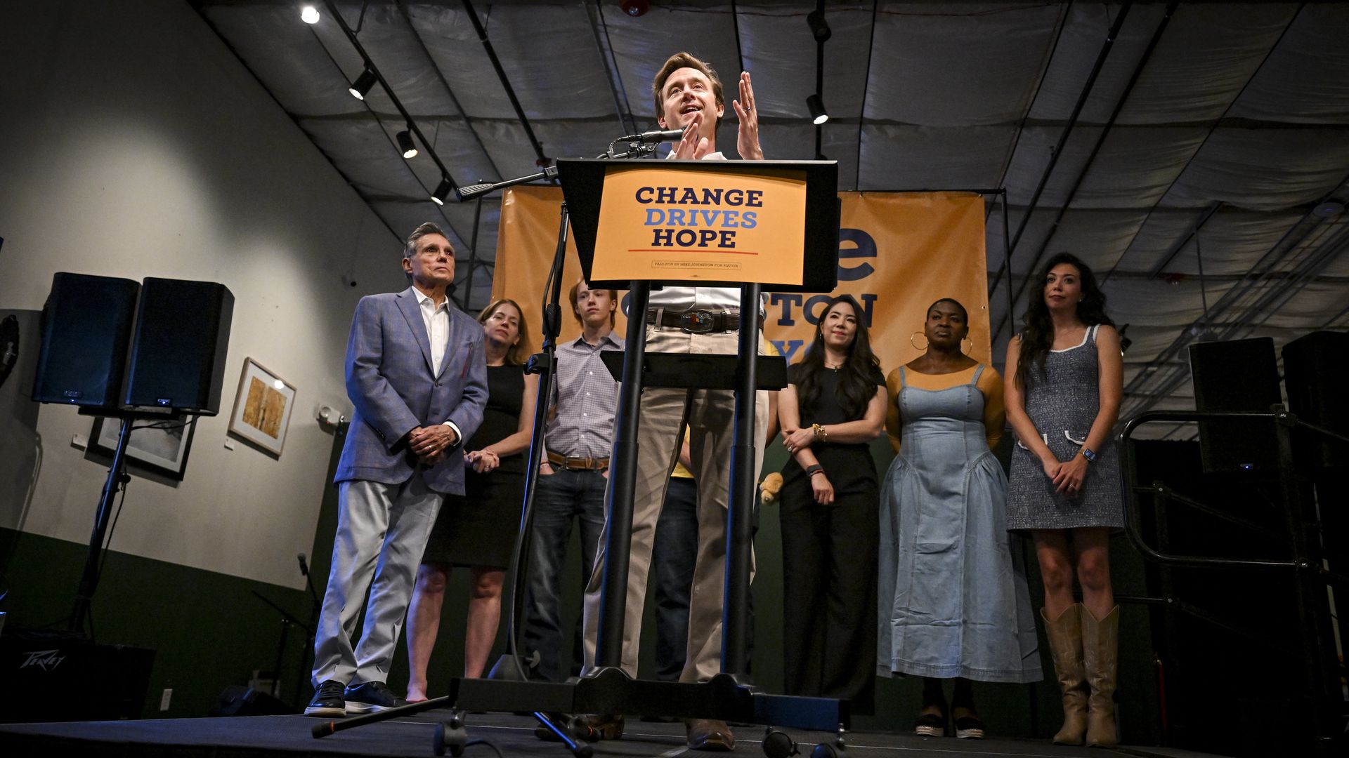 A photo of Mayor Mike Johnston standing on stage in front of a podium that has a campaign sign reading "Change drives hope" with a half-dozen people standing behind him.
