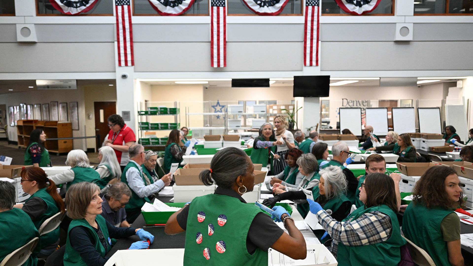 Busy indoor hall with volunteers in green vests at long tables sorting papers; American flags and red-white-blue bunting hang from the upper level as people sort envelopes.