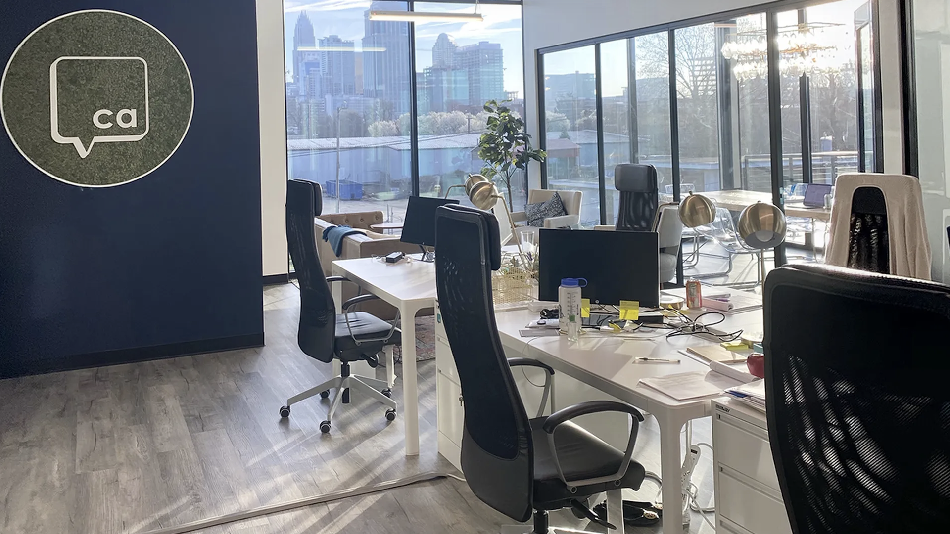 Open-plan office with white desks and black ergonomic chairs, large glass walls overlook a city skyline. A dark blue wall displays a round logo with CA inside a speech-bubble.