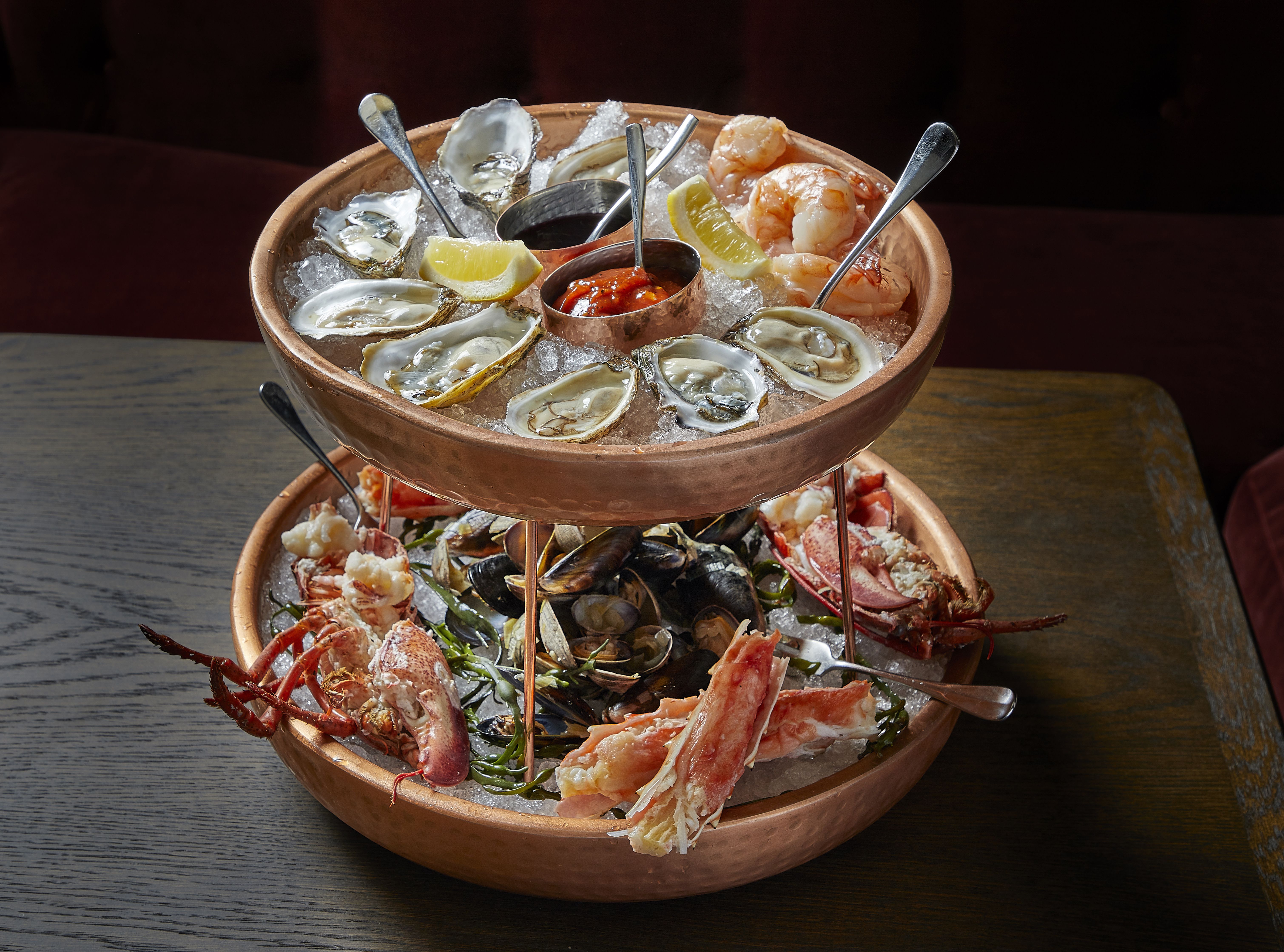 A double layer seafood tower with oysters on the top bowl and shellfish on the bottom.