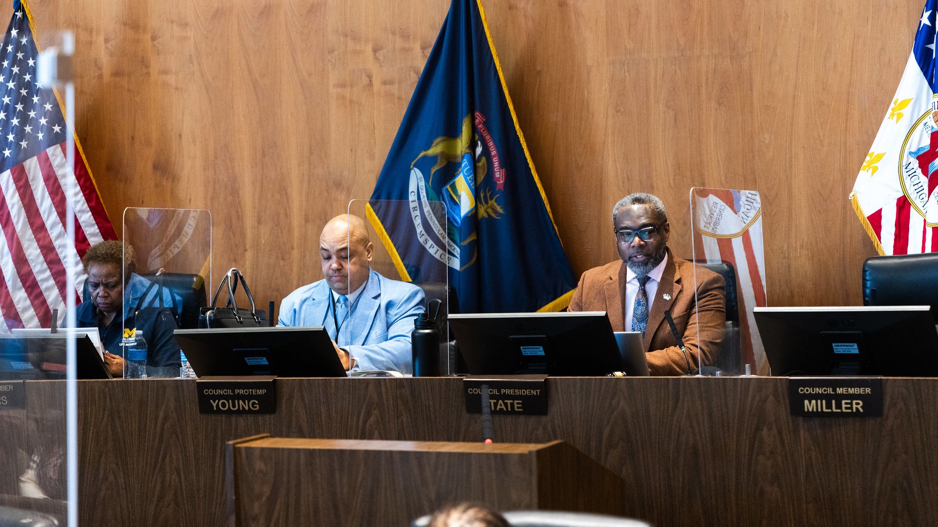 Three city council members sit at their desks looking out at the auditorium 