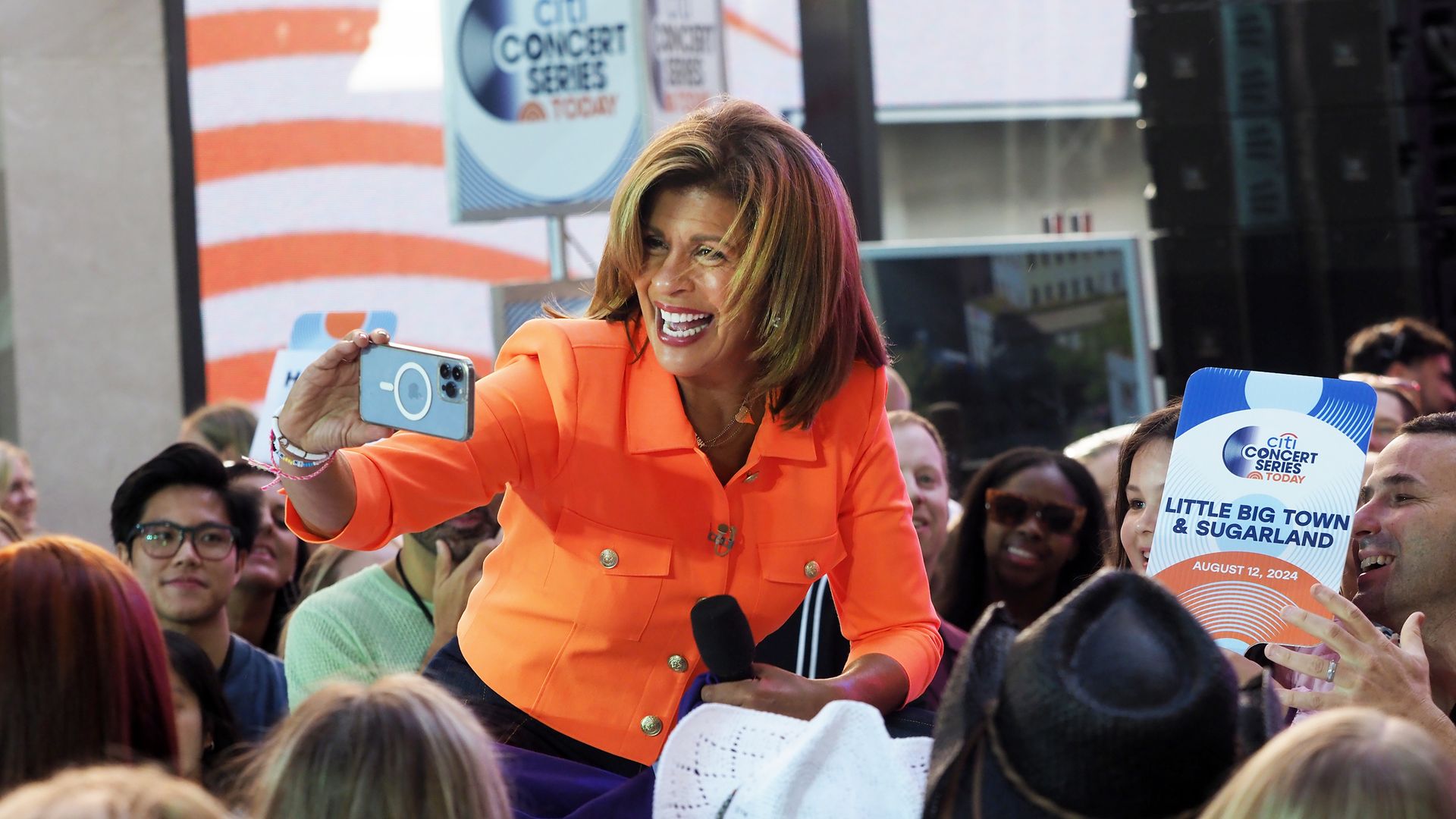 A person in an orange suit jacket takes a selfie amid a crowd while holding a microphone