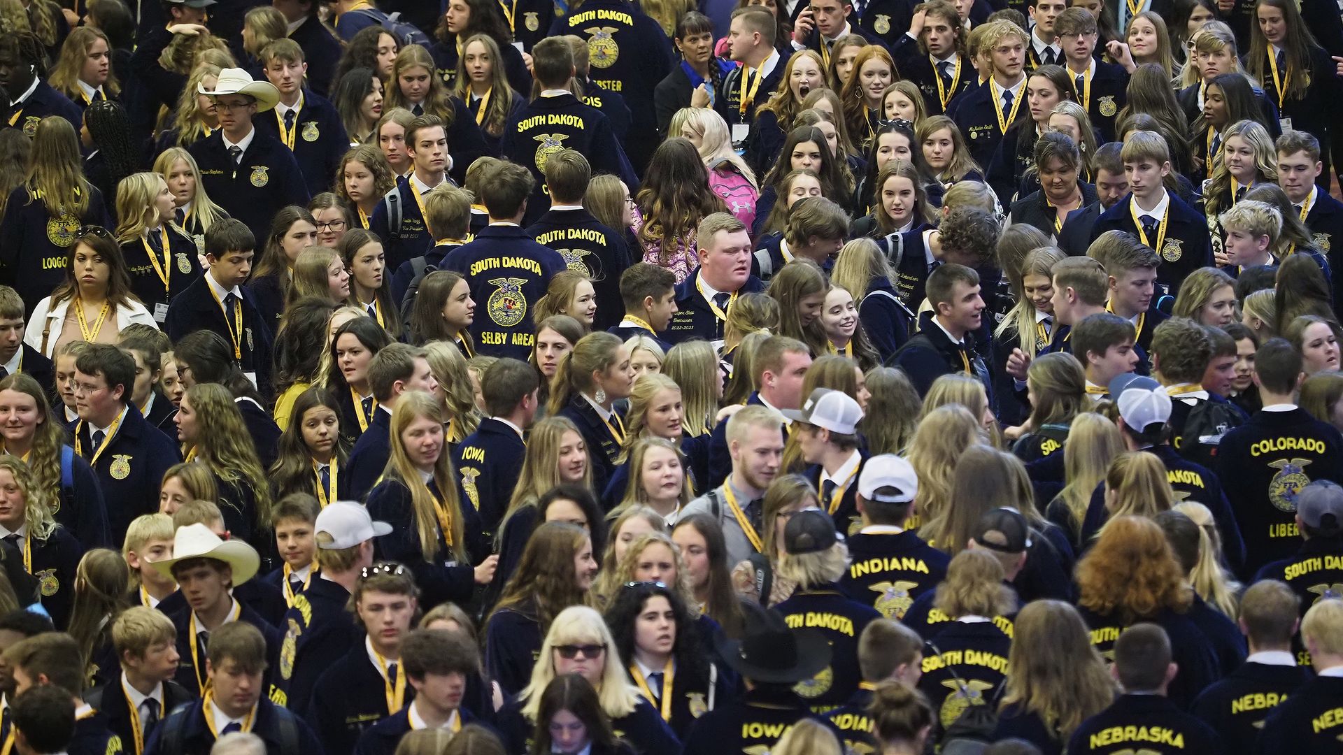 Large crowd of students in navy blue FFA jackets with gold embroidery and yellow lanyards; jackets display state names such as South Dakota, Indiana, and Nebraska as they mingle at a busy event.