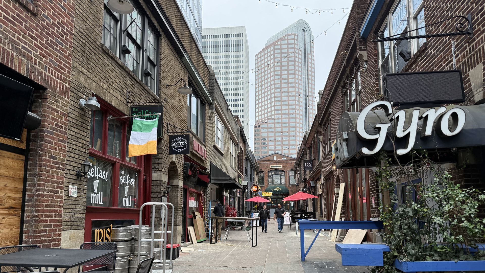 Urban alley with brick buildings and outdoor seating, hanging Irish flag, signs for "Gyro" and "The Belfast Mill," two people walking, and tall modern buildings in the background under a gray sky.